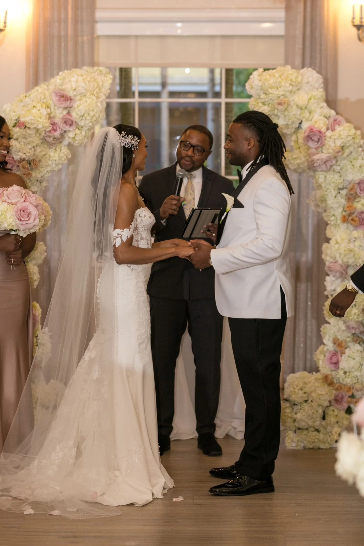 A wedding ceremony with the bride and groom holding hands and facing each other, surrounded by floral arrangements, with The Florida Pastor standing with them, in a decorated indoor venue.