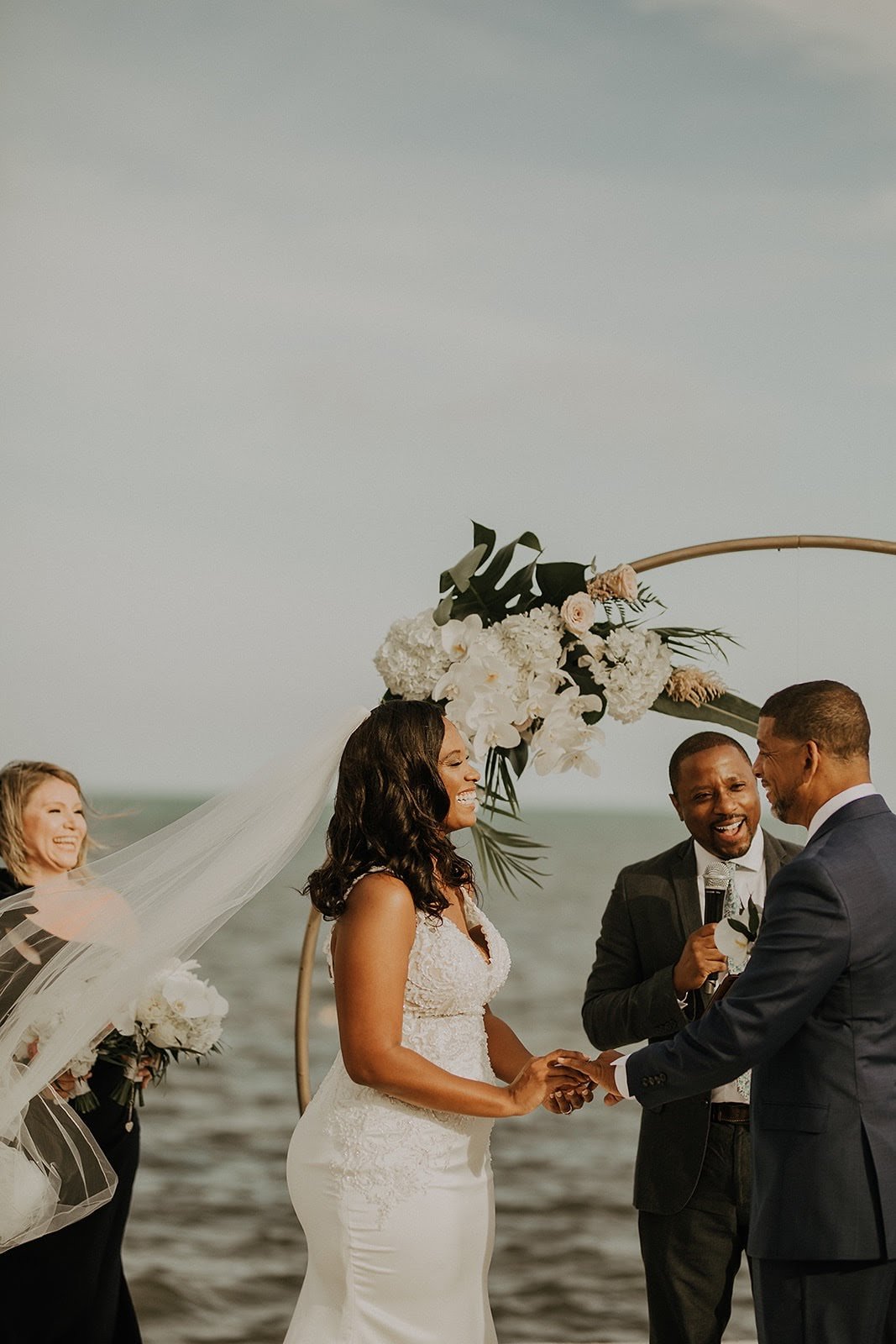A couple exchanging wedding vows at the beach with The Florida Pastor and bridesmaid present, under a floral arch.