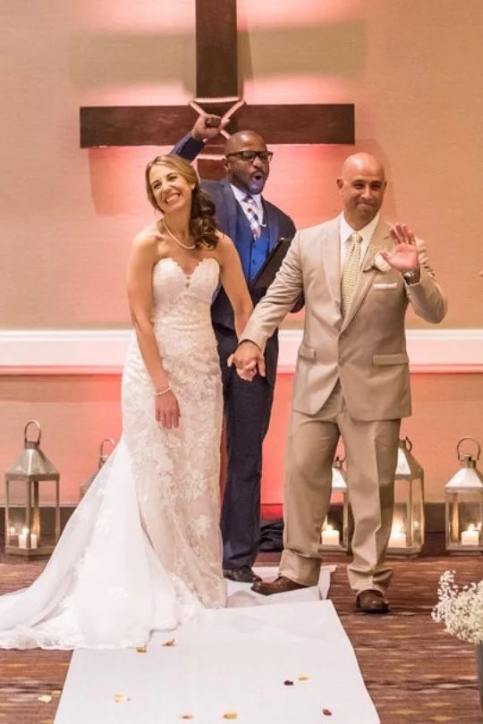 A wedding ceremony with a bride in a white lace dress, a groom in a beige suit, and The Florida Pastor with his hand behind a cross in a church setting with lanterns and candles.