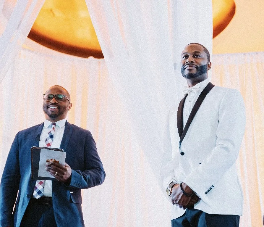 The Florida Pastor and groom standing on a stage with white curtains and a yellow overhead structure,  both smiling.