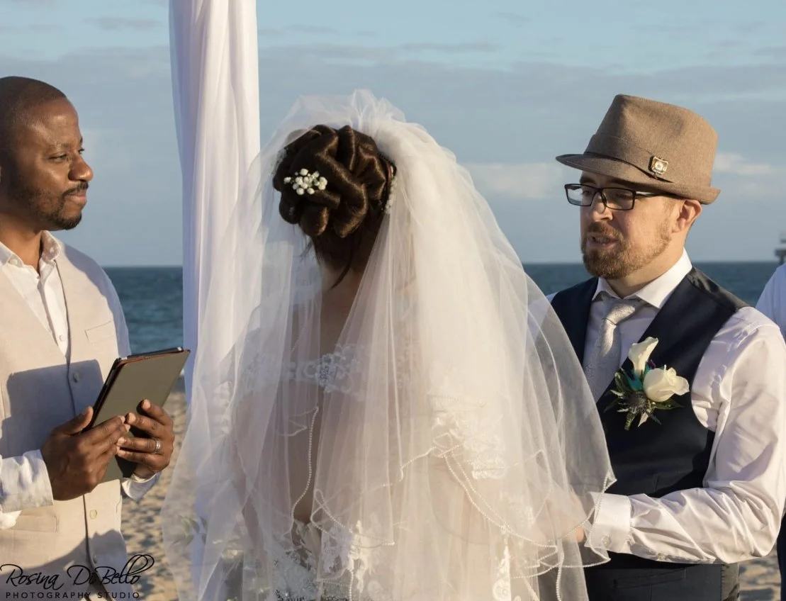 A wedding ceremony officiated by The Florida Pastor, taking place on the beach during sunset, with a bride in a white wedding dress and veil, a groom in a vest and tie.