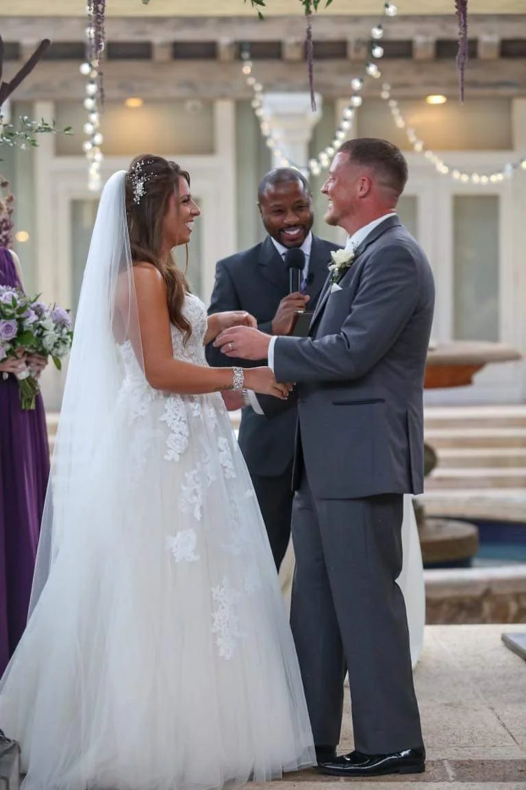 A bride and groom exchanging vows at their wedding ceremony, with The Florida Pastor holding a microphone behind them, in an outdoor setting decorated with string lights.