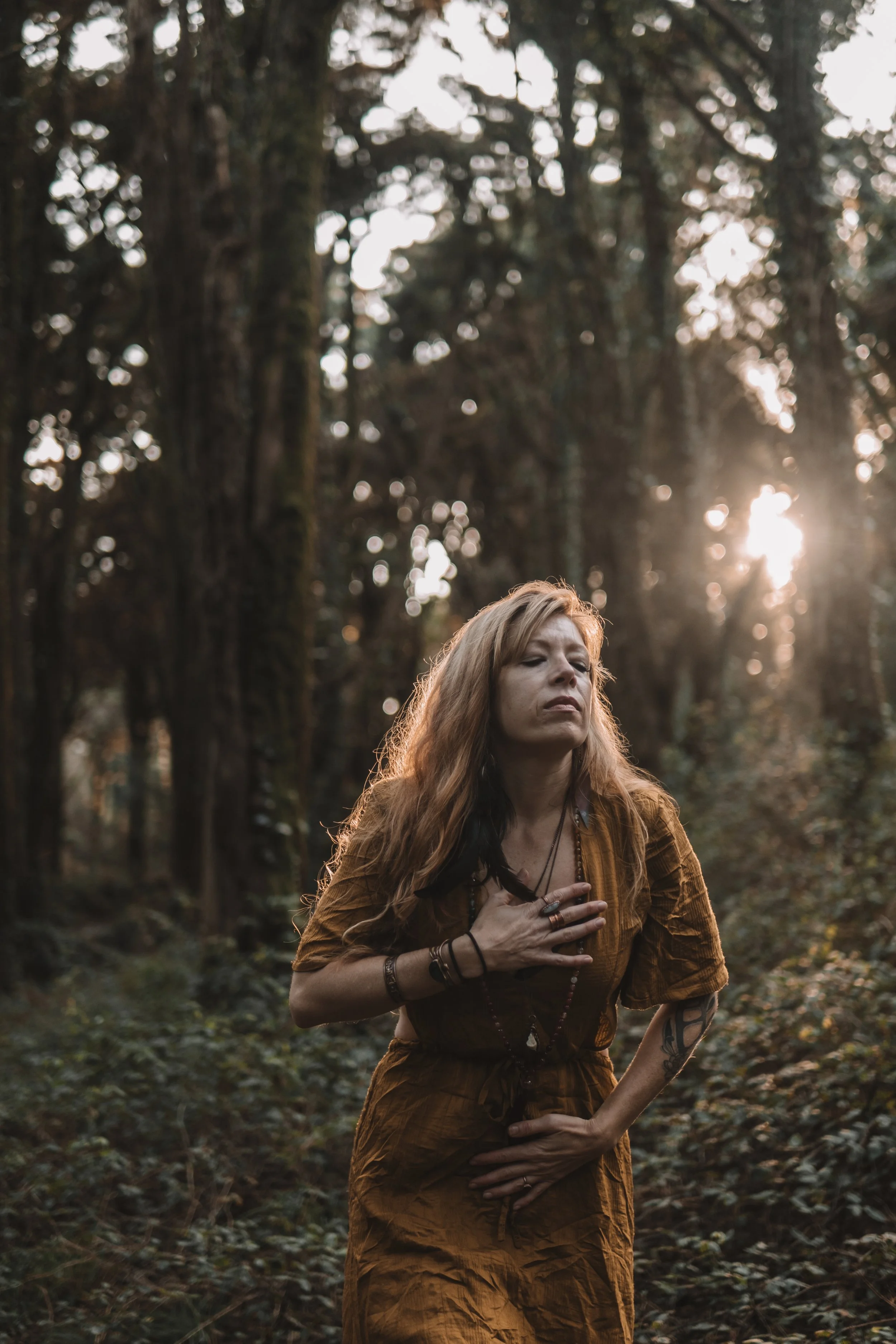 A woman with long wavy hair standing in a wooded forest during sunset, wearing a brown dress with jewelry and tattoos, with her hand over her chest.