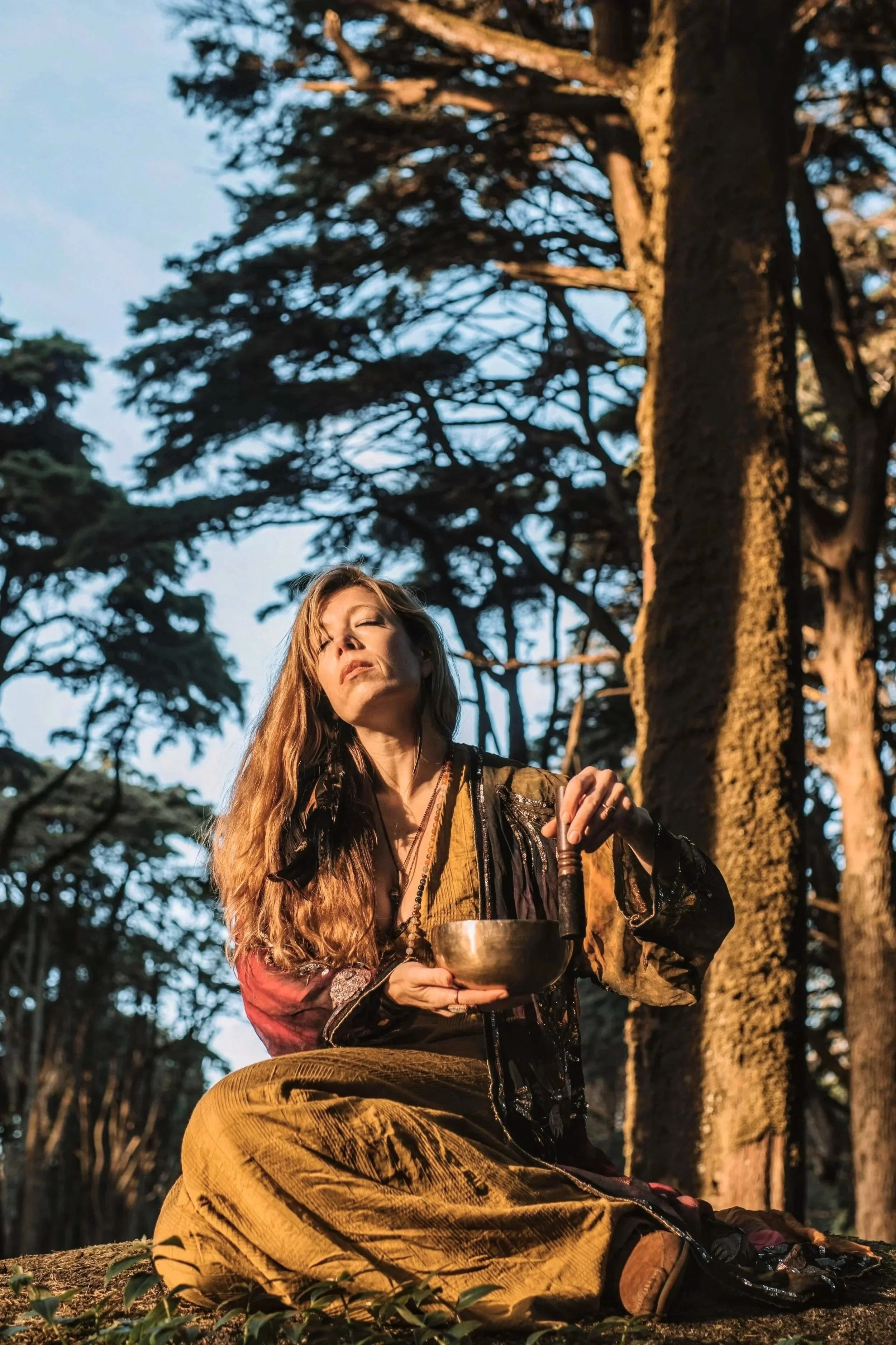 A woman with long red hair sitting cross-legged outdoors in a forest, holding a singing bowl and a wooden mallet, with eyes closed and a serene expression, illuminated by warm sunlight.