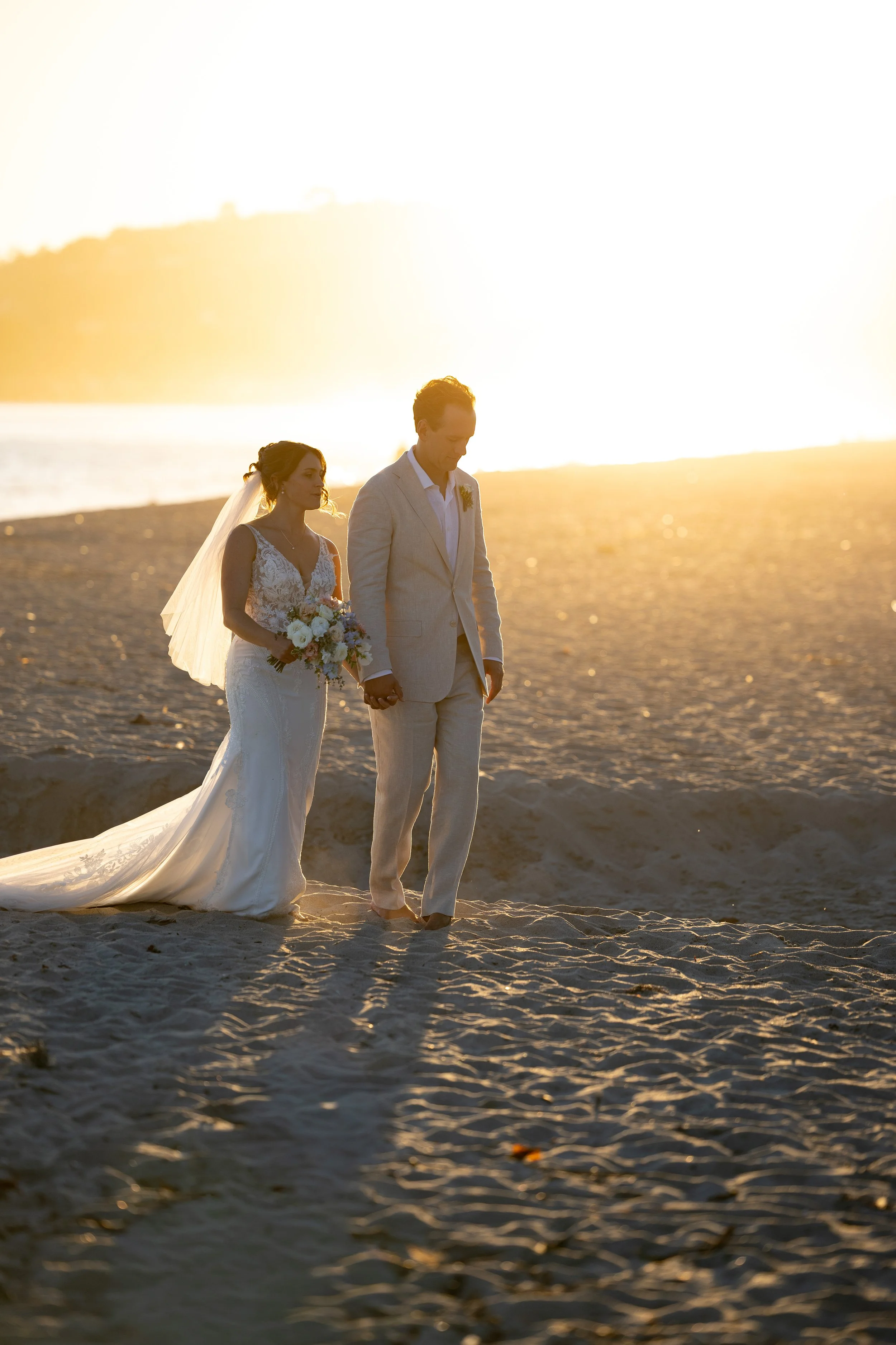 A bride and groom holding hands on the beach during sunset.