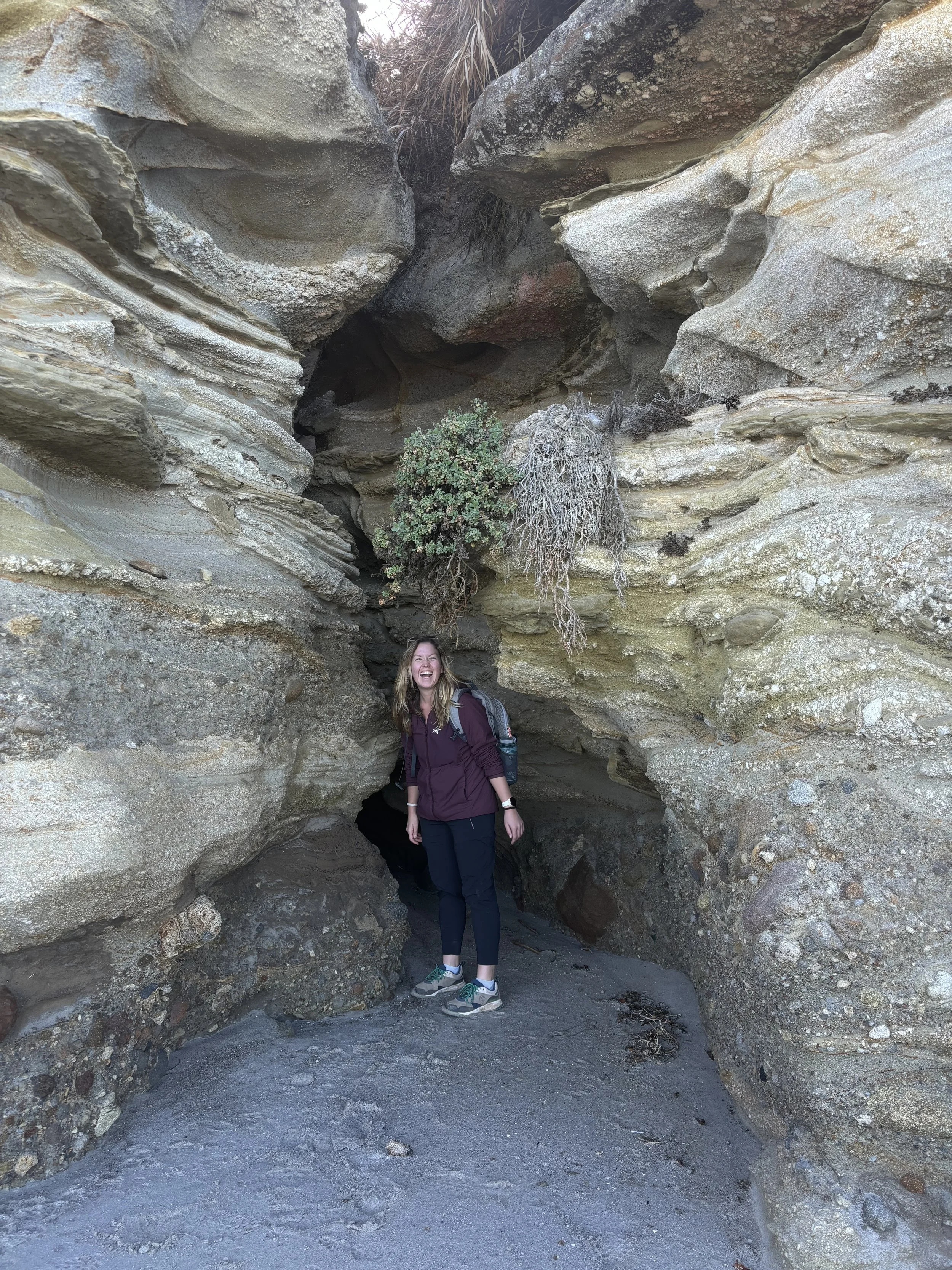 A woman smiling and standing inside a narrow slot canyon with rugged, layered rock walls and a small, sparse green bush above her.