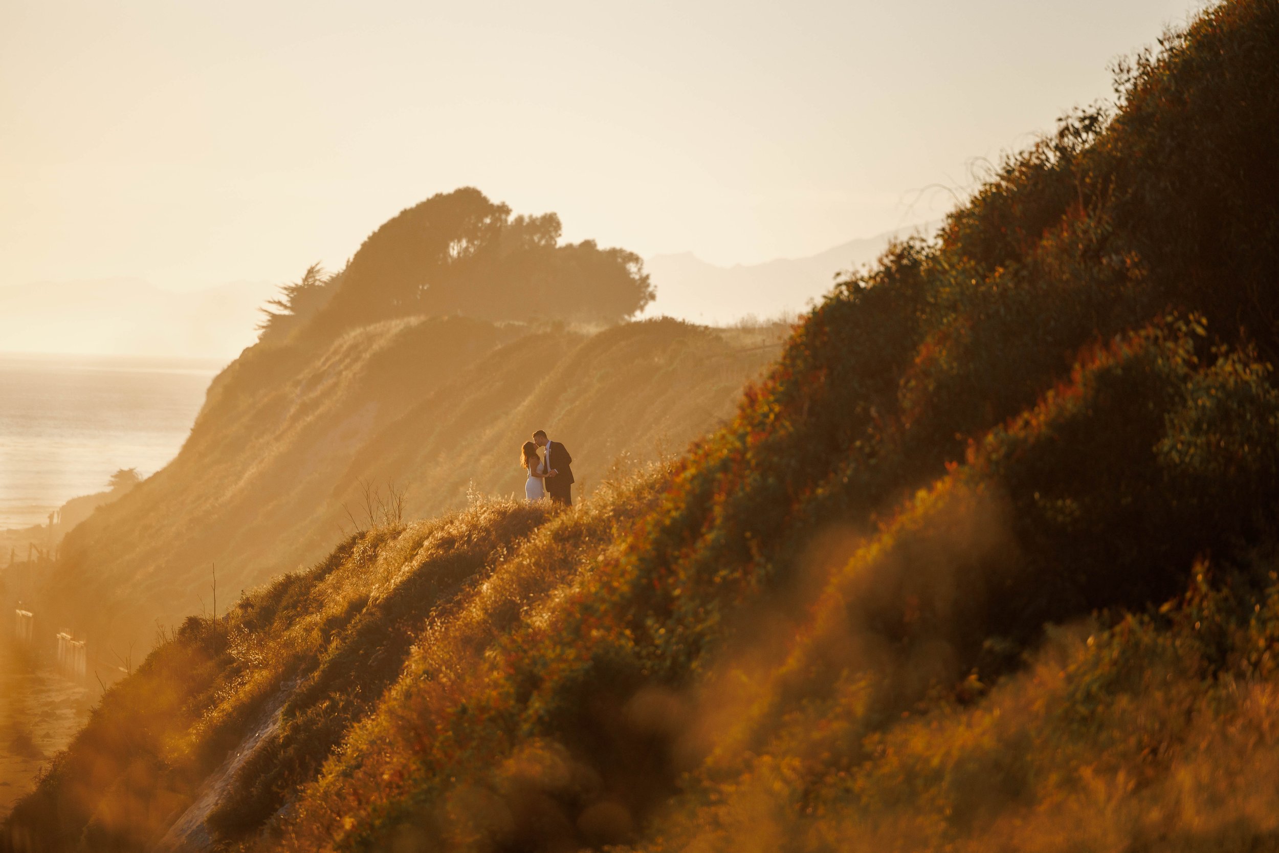 A couple standing close together on a hillside at sunset, with a scenic view of rolling hills and the ocean in the background.