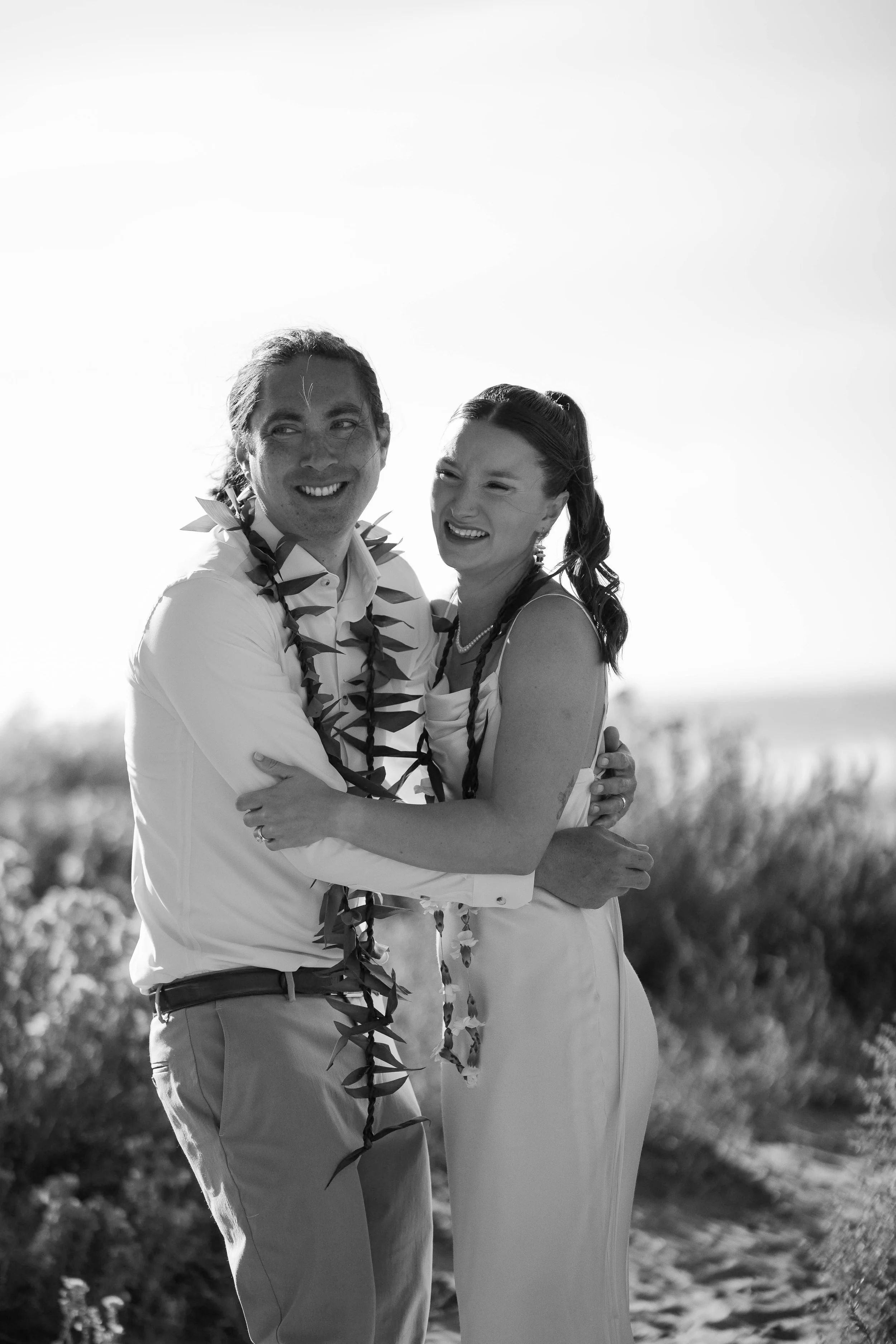 A black and white photo of a smiling couple embracing outdoors, wearing leis, on a sunny day with blurred shrubbery in the background.