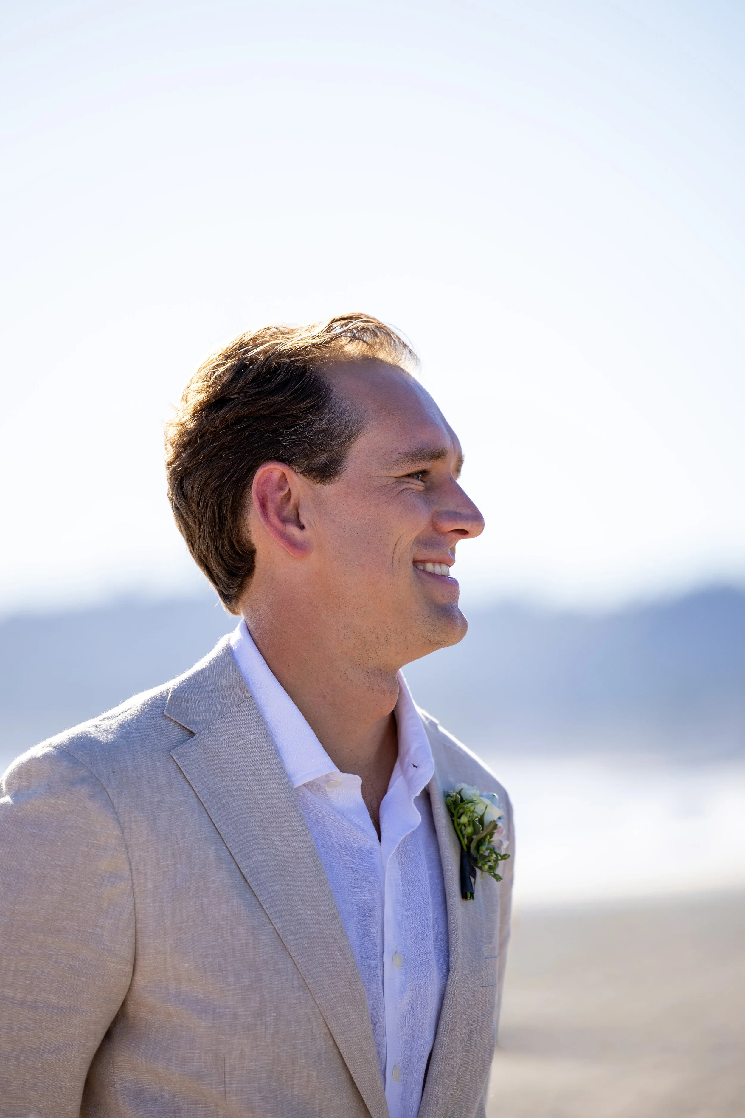 A man dressed in a light-colored suit with a boutonniere, smiling while looking to the side outdoors with a bright sky and distant mountains in the background.