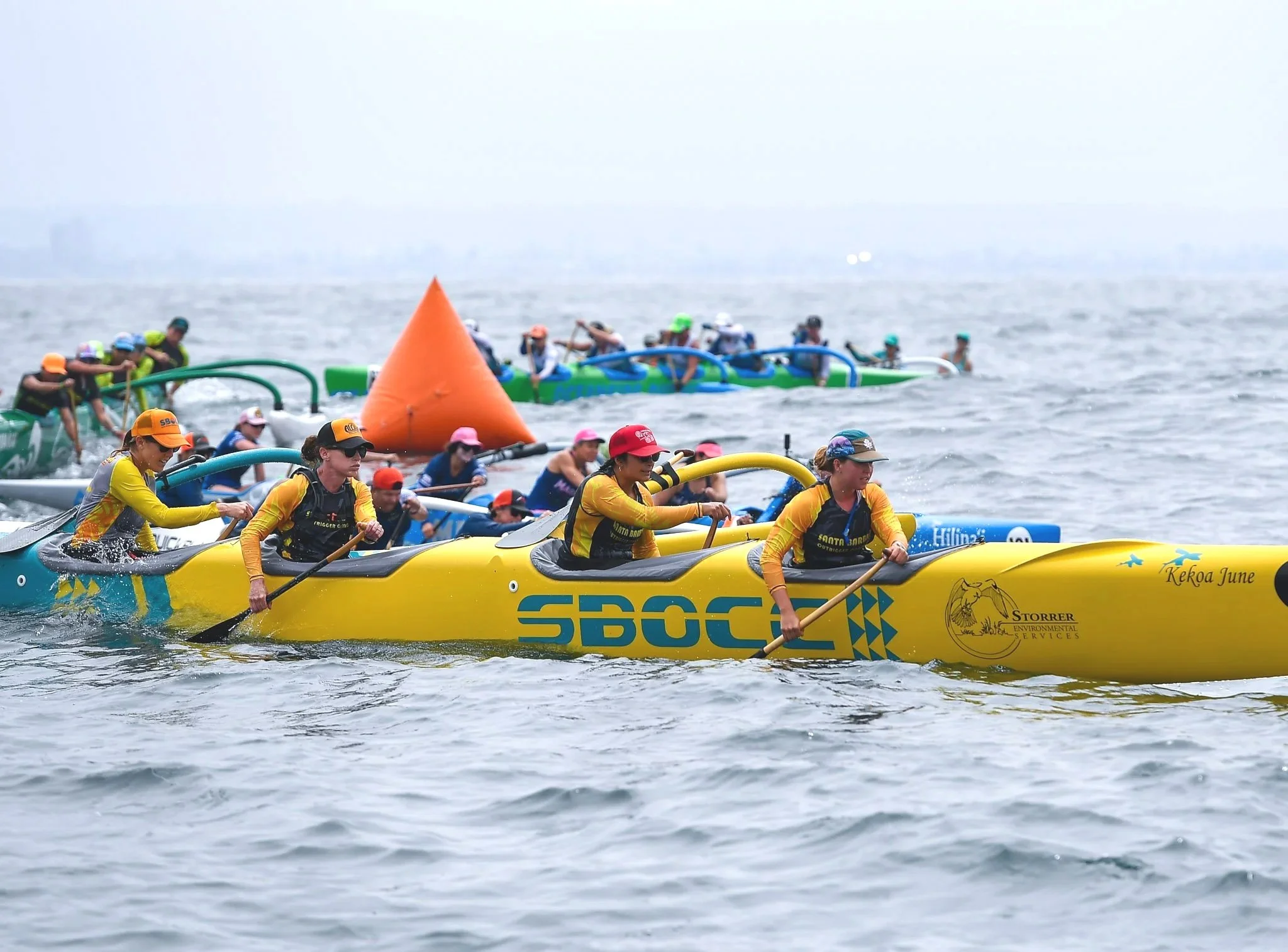 A group of women competing in a canoe race on open water, paddling in yellow and blue canoes near a large orange buoy, with other teams and boats visible in the background.