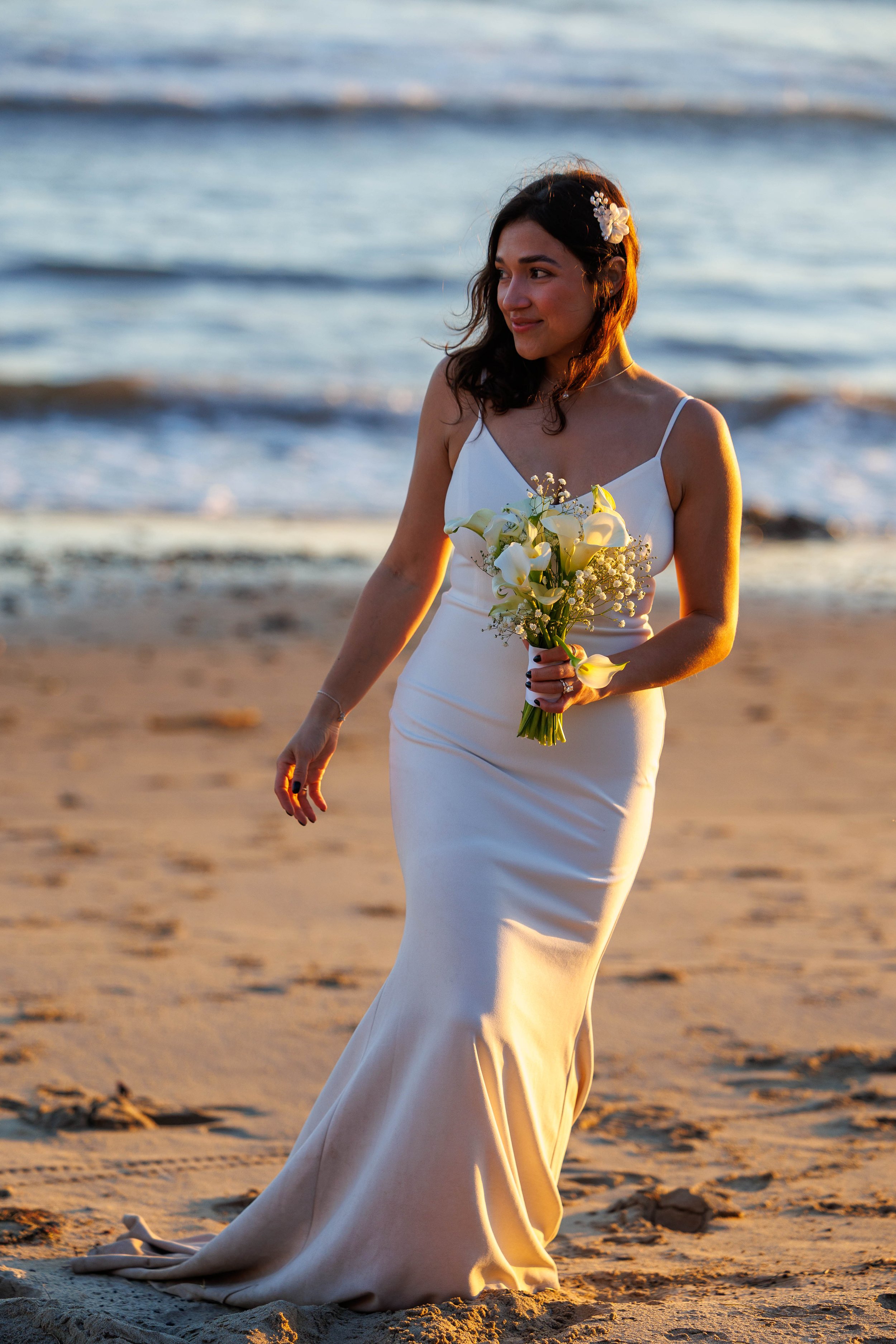 A woman in a white wedding dress holding a bouquet of white flowers walking on the beach at sunset.