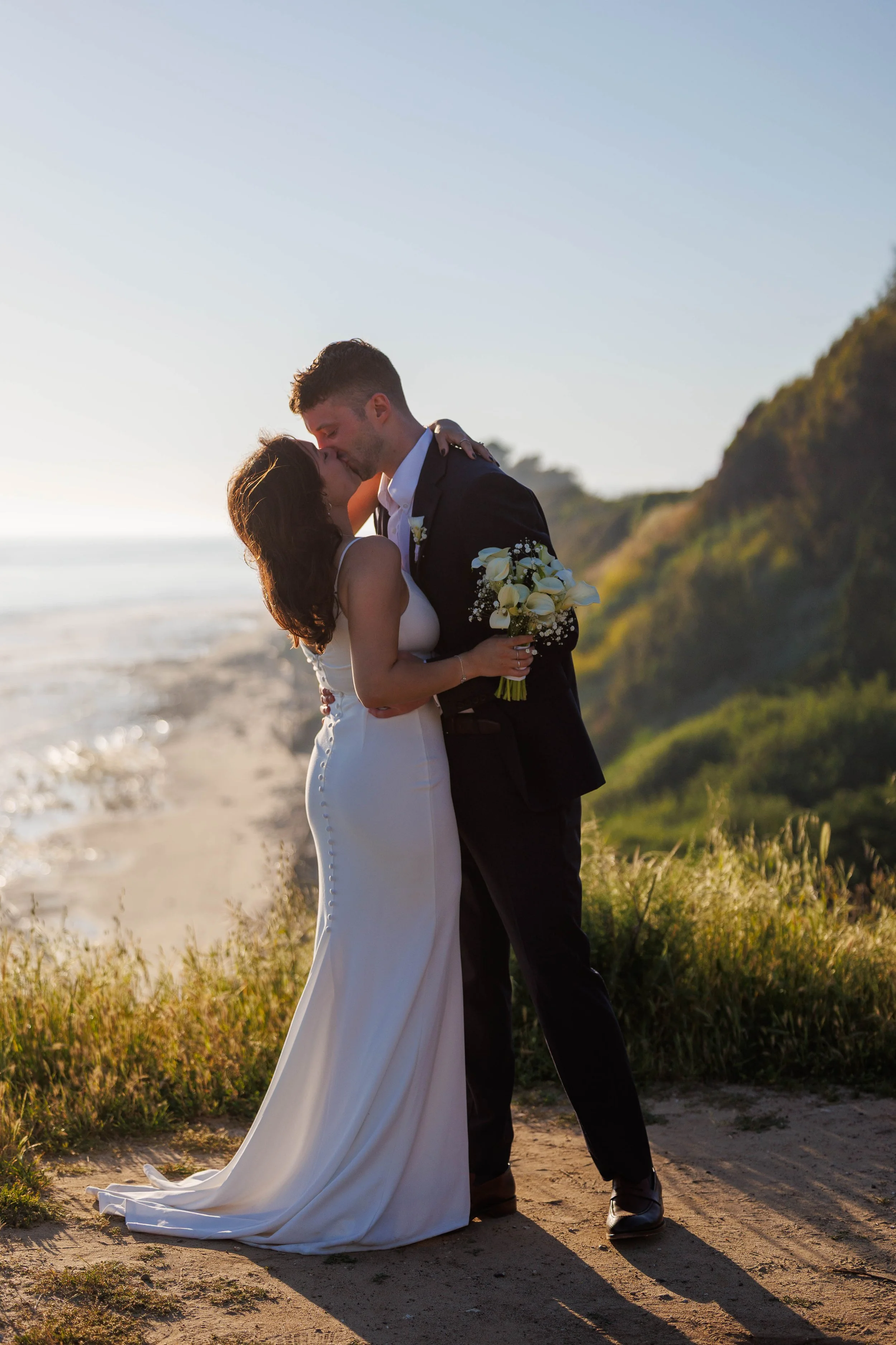 A newlywed couple sharing a kiss on a beach at sunset, with greenery and cliffs in the background.
