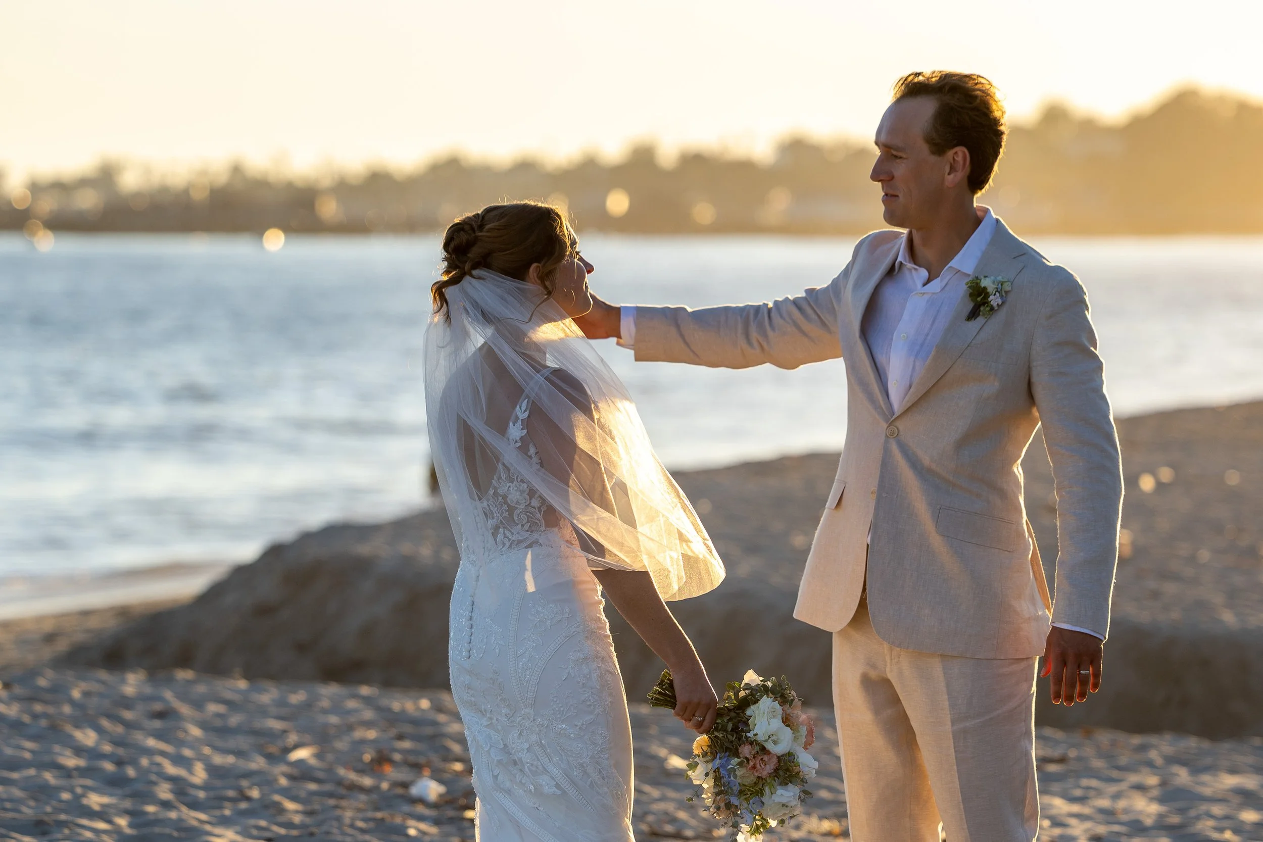 A bride and groom standing on a beach at sunset, with the groom touching the bride's face while she holds a bouquet of flowers.