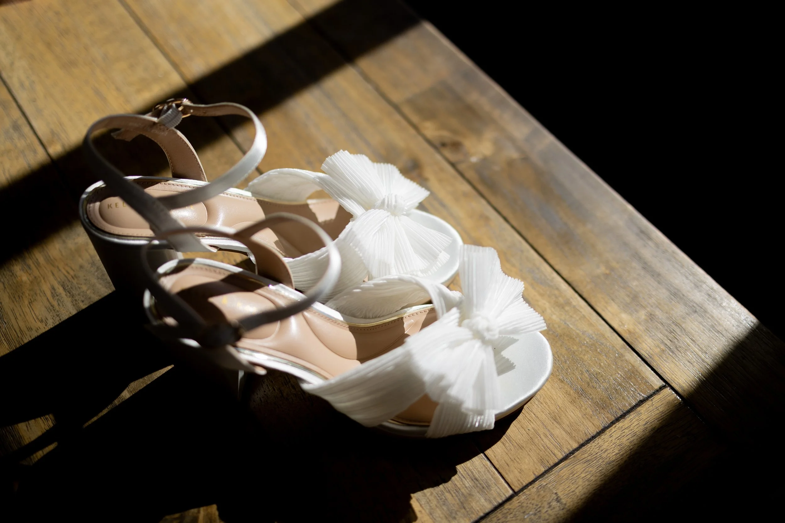 A pair of beige high-heeled shoes with ankle straps, adorned with white fabric flowers, placed on a wooden floor with sunlight casting a shadow.