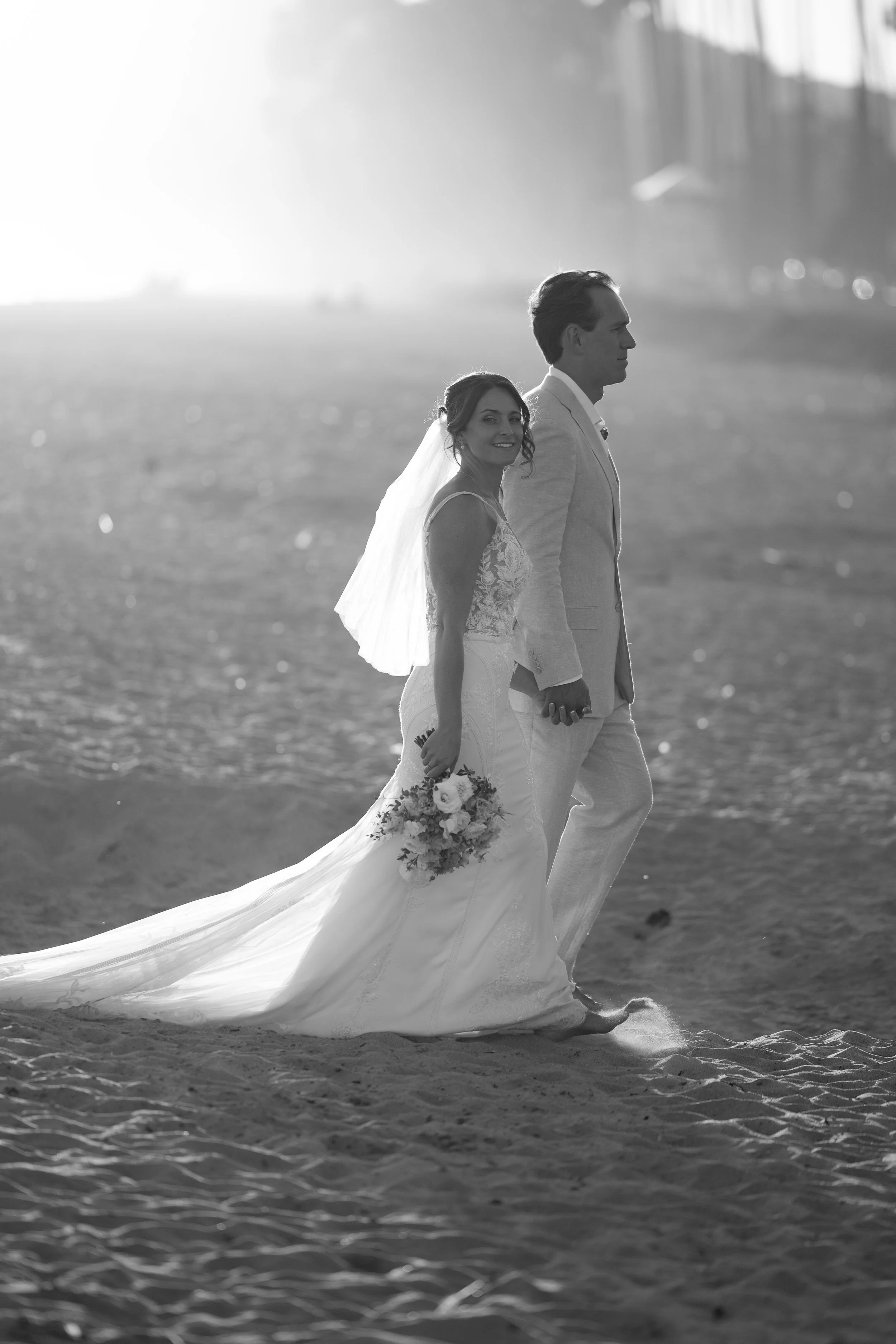 A black-and-white photo of a newlywed couple walking on the beach holding hands, with the bride smiling and holding a bouquet of flowers, and the groom looking forward.
