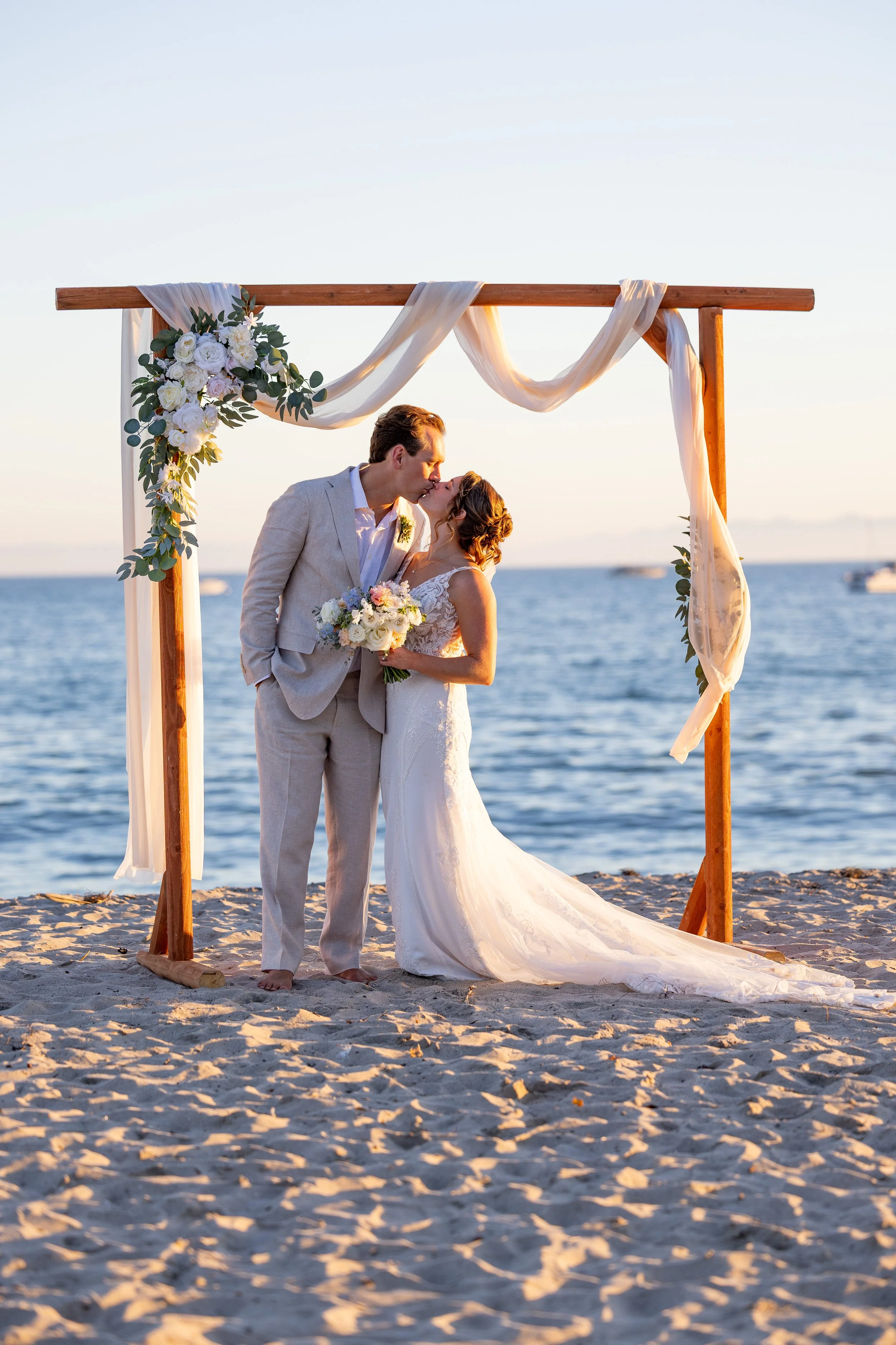 A newlywed couple sharing a kiss under a wooden wedding arch decorated with flowers and draped fabric on a beach at sunset.