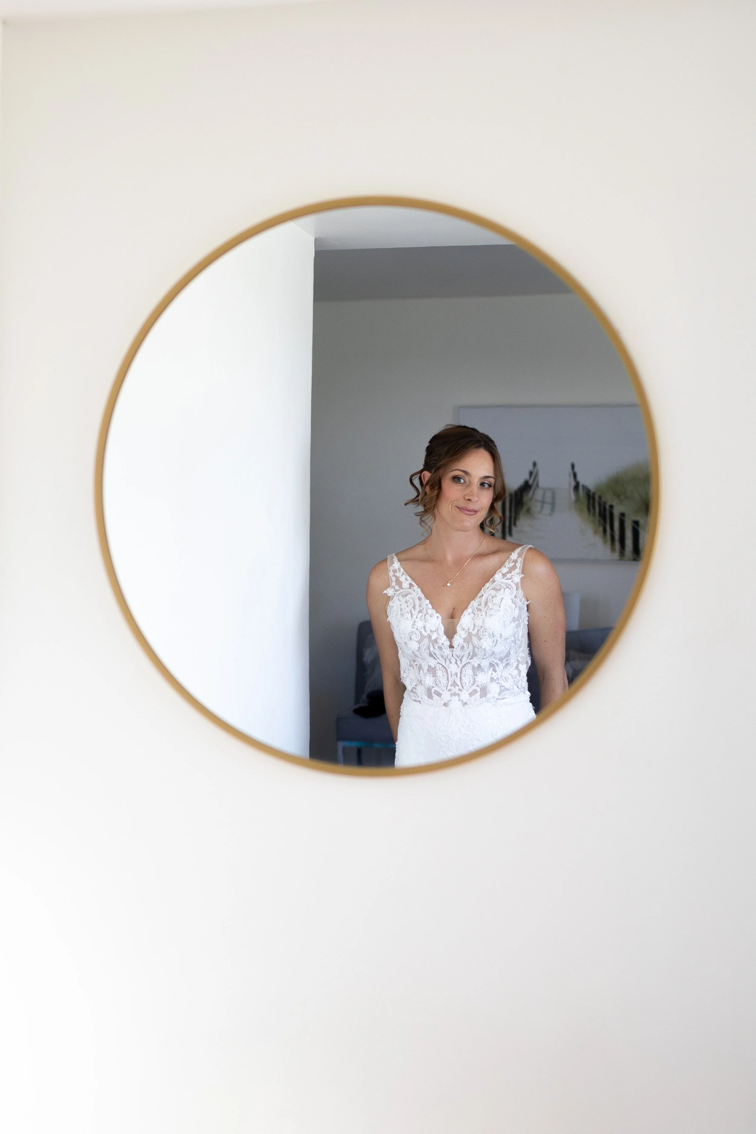 A woman in a white lace dress is seen through a round mirror, standing in a room with a simple background and a wall painting of a bridge over water.