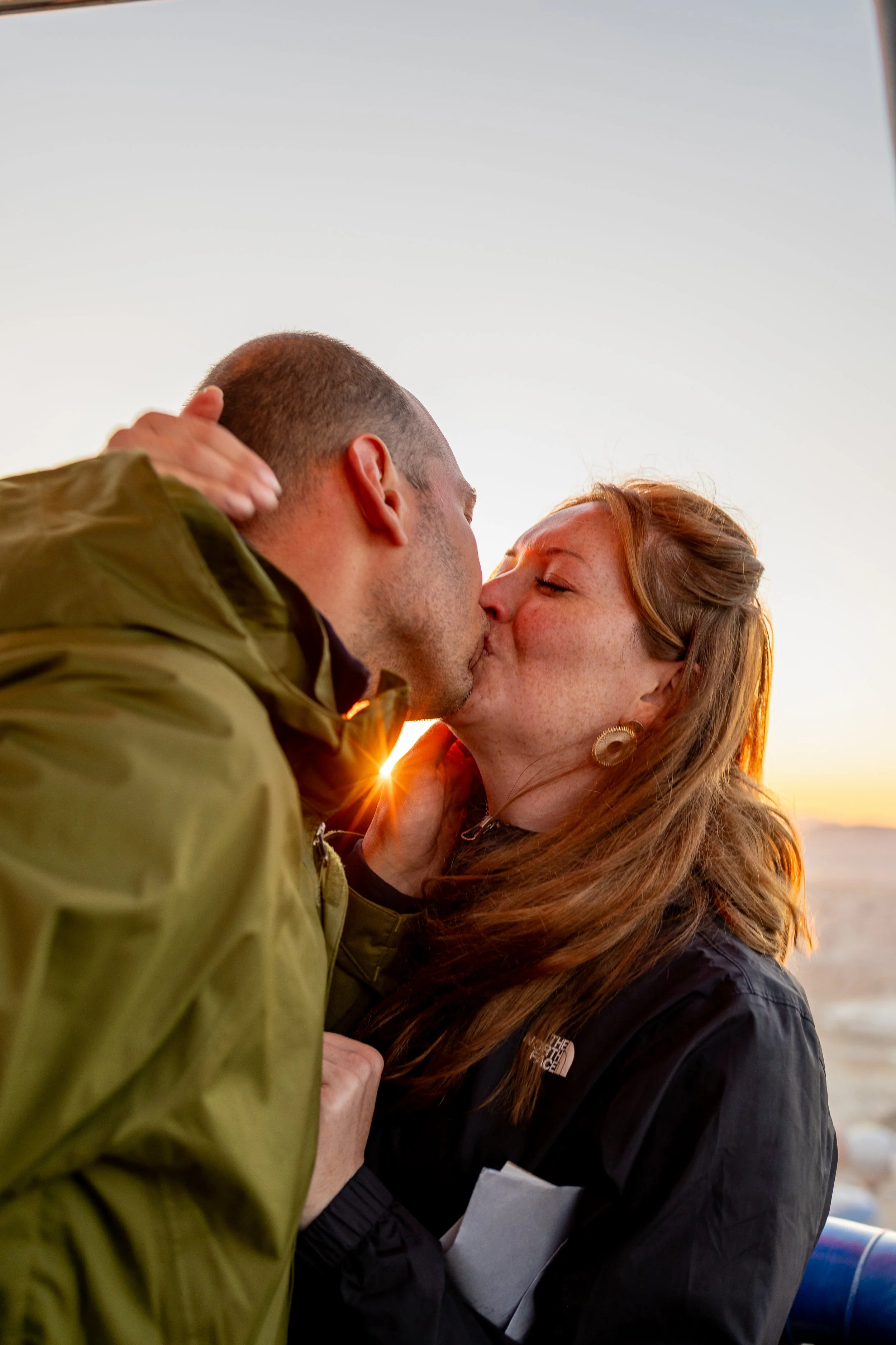 A couple sharing a kiss outdoors at sunset, with the sun partially visible behind them.