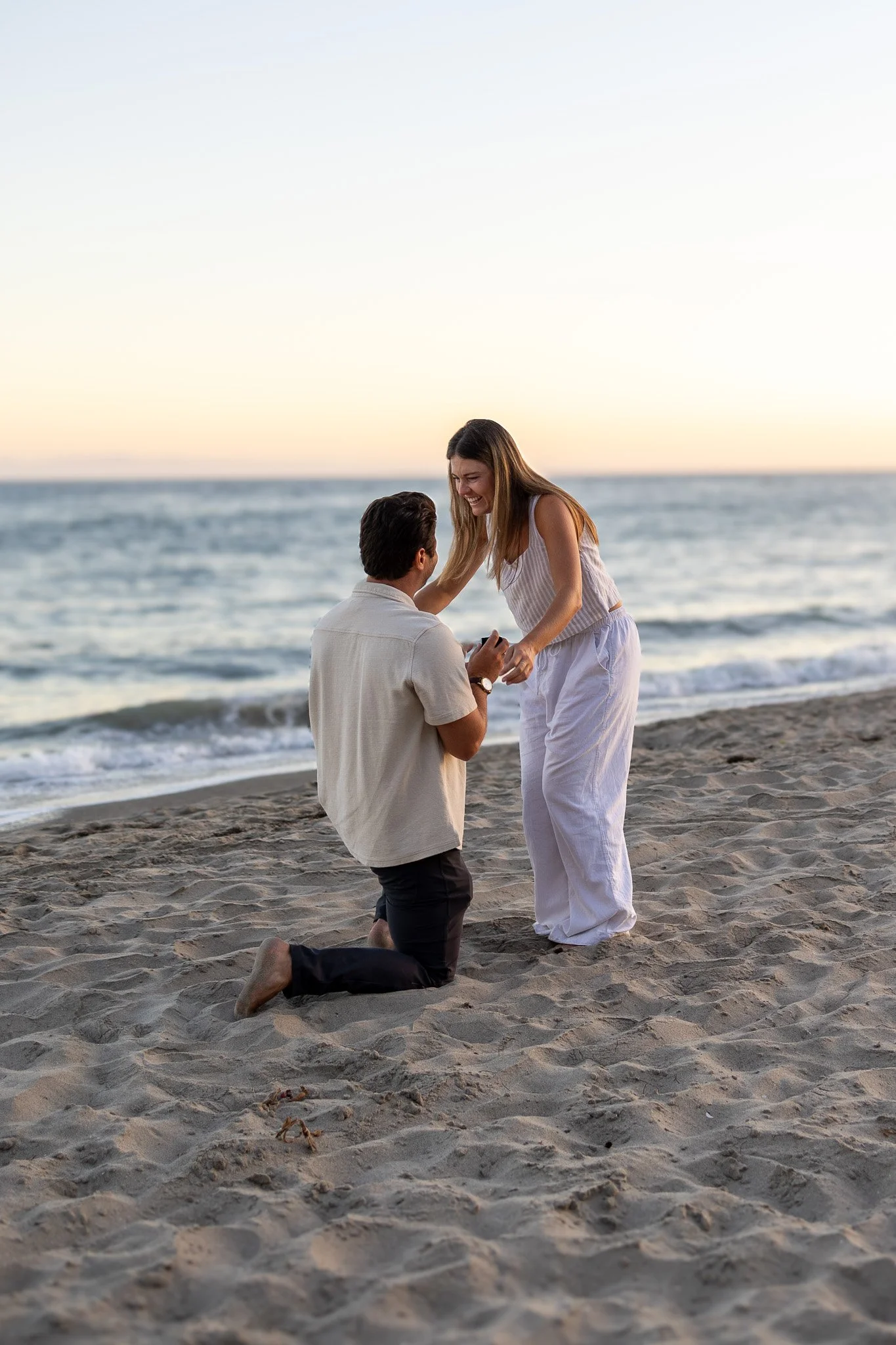 A couple having a marriage proposal on the beach at sunset, with the man kneeling and the woman smiling.