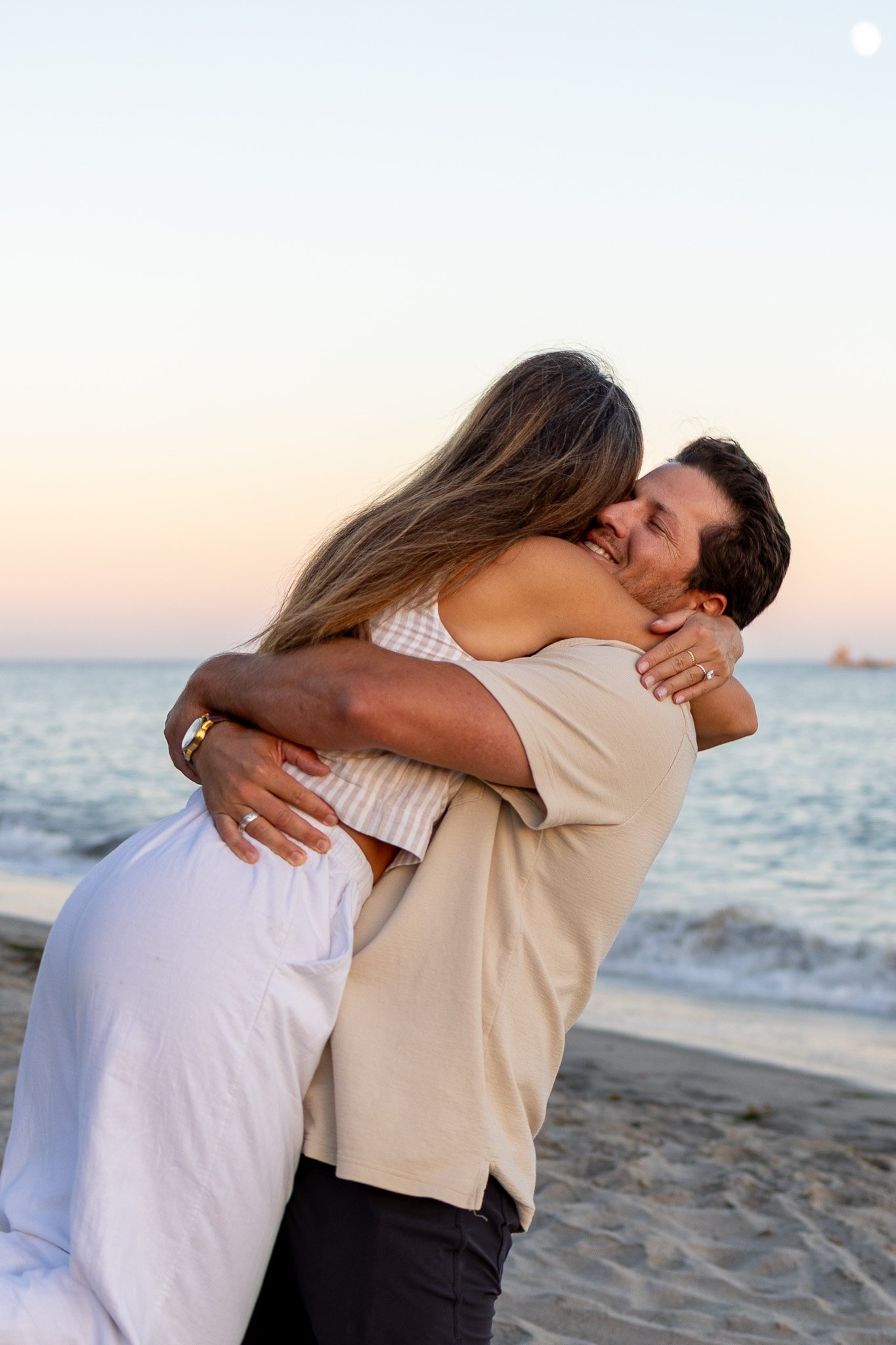 A couple hugging on the beach during sunset, smiling happily.