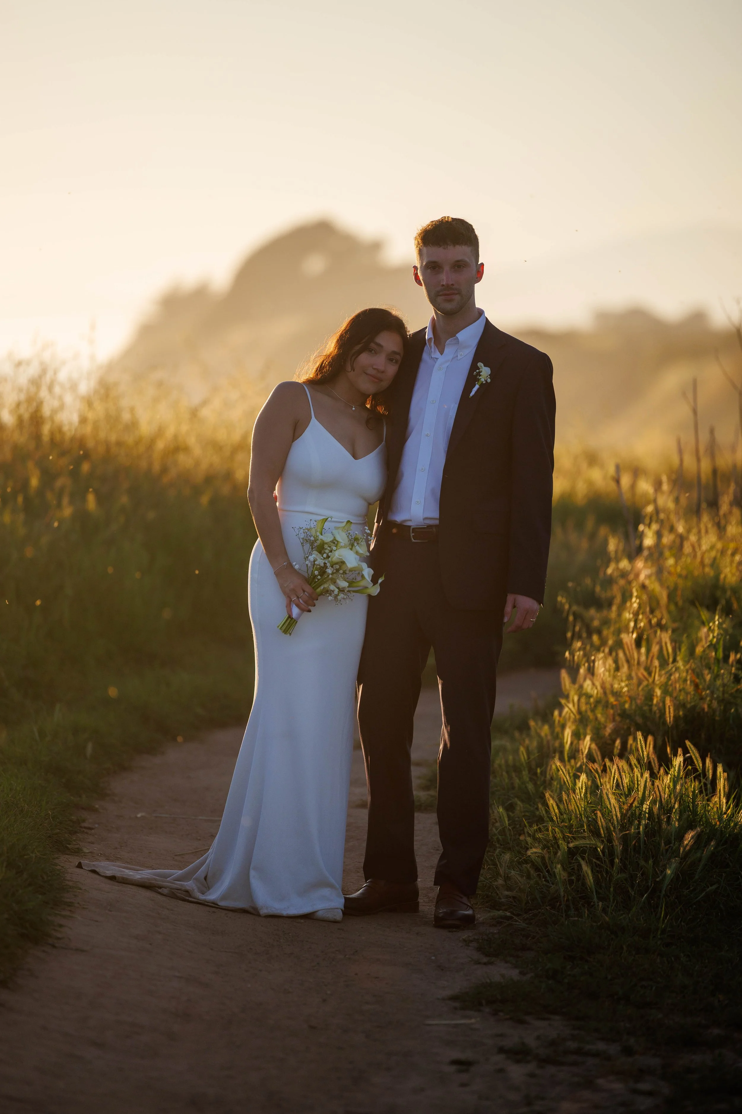 A bride and groom standing together outdoors during sunset, with the bride holding a bouquet and the groom wearing a suit and boutonniere.