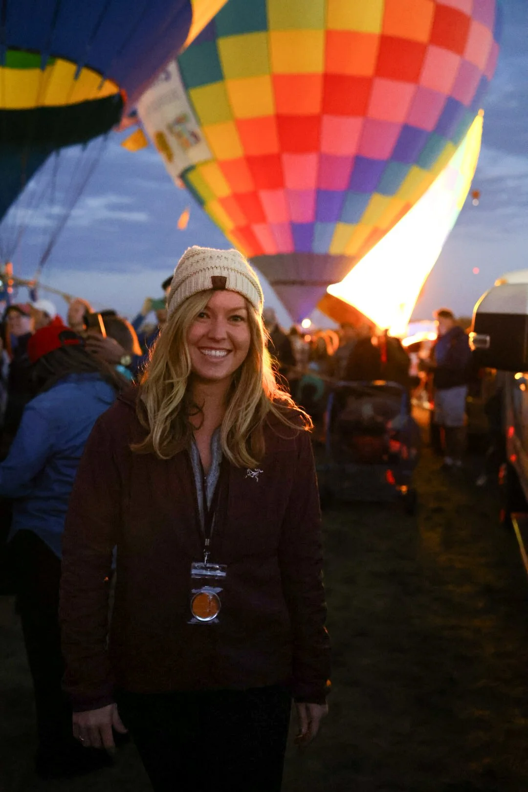 A woman smiling at a hot air balloon festival during dusk, with colorful balloons filling the sky behind her.