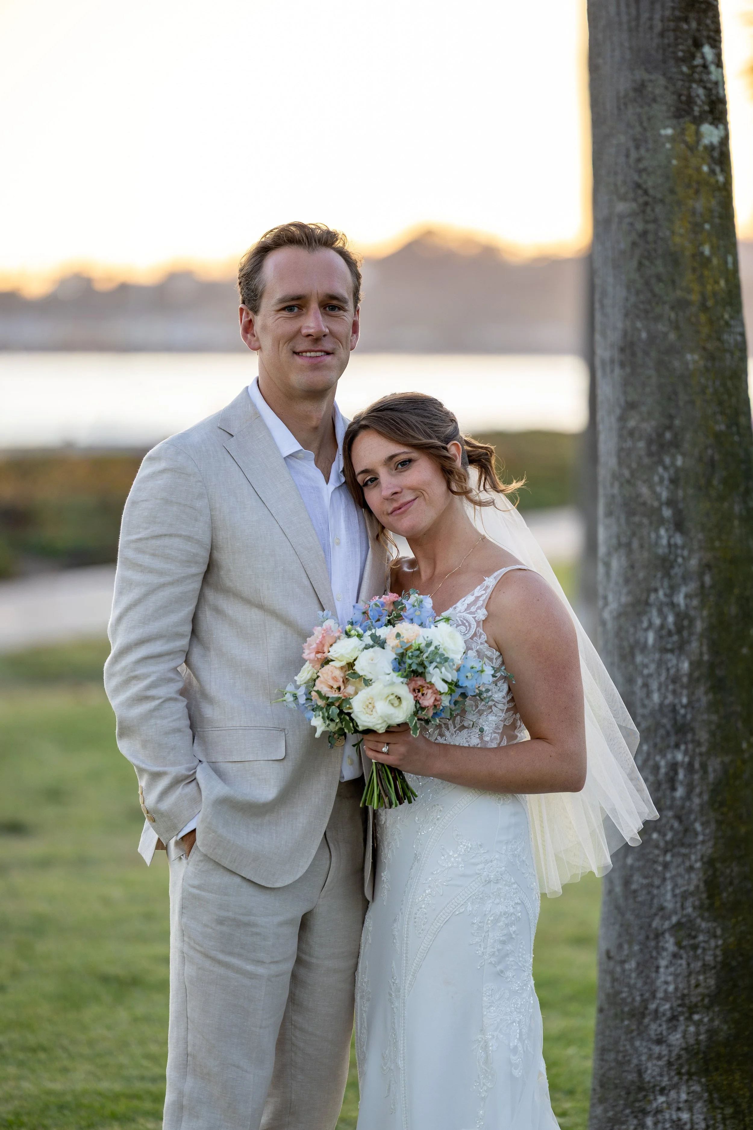 A bride and groom standing outdoors near a tree during sunset, with the bride holding a bouquet of flowers and both smiling.