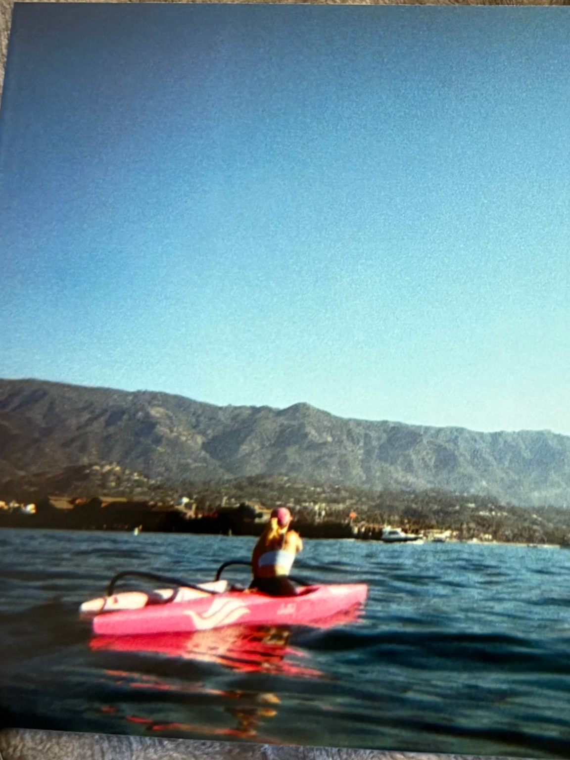 A person kayaking on a lake or ocean, with mountains in the background and clear blue sky overhead.