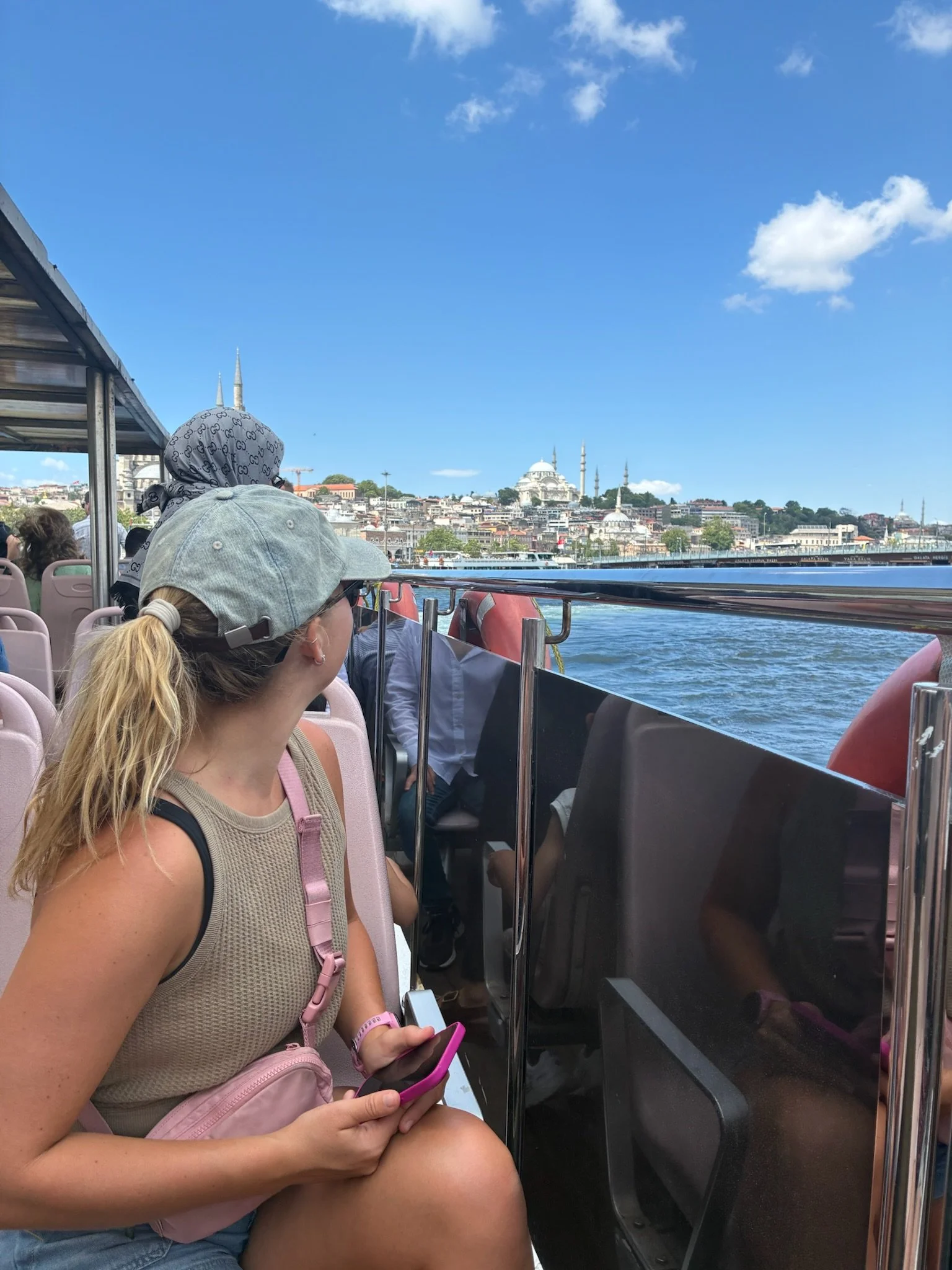 A woman with a gray baseball cap and beige tank top sitting on a boat, holding a pink phone, looking towards the city skyline with mosques and minarets across the water.