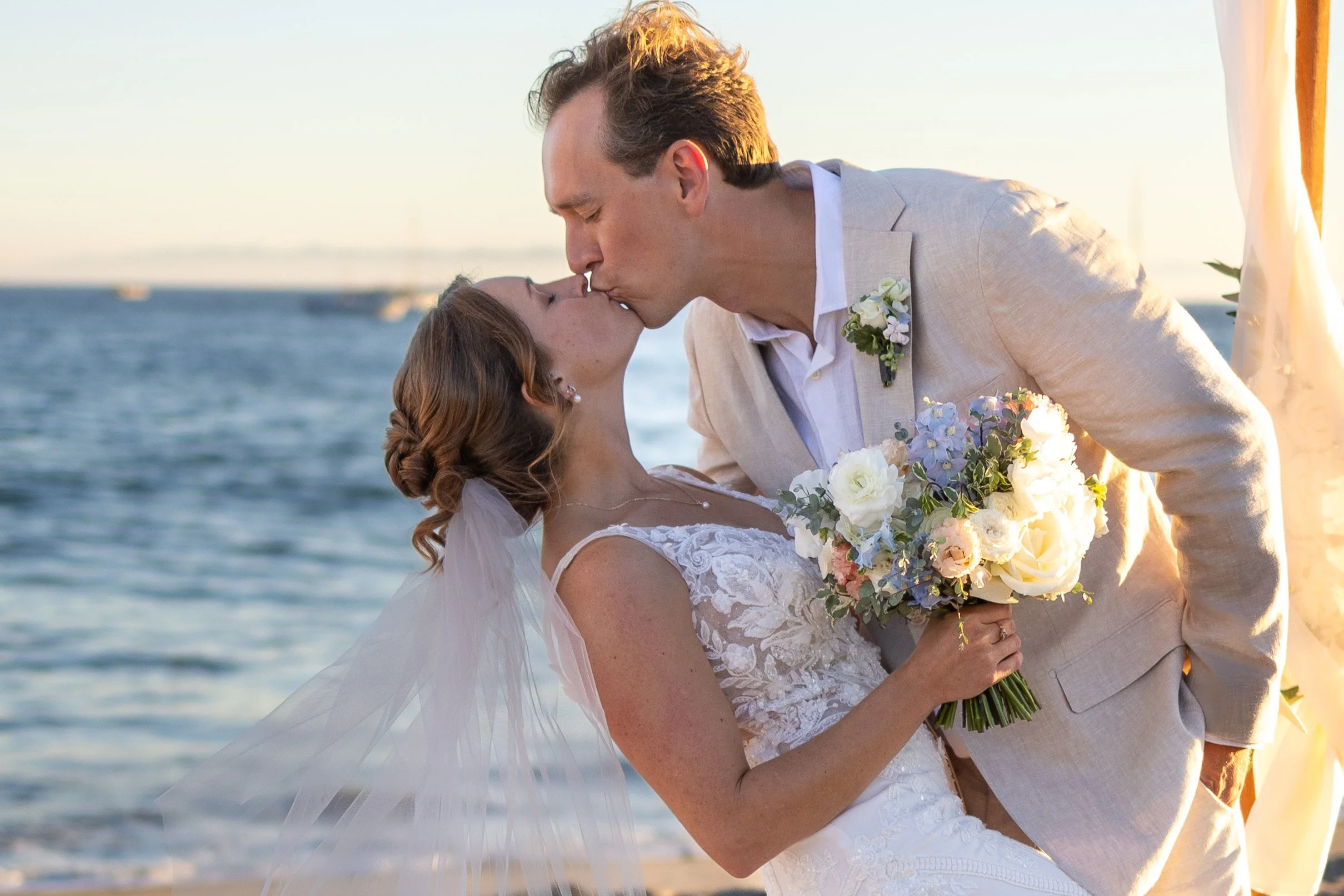 A bride and groom share a kiss at a beach wedding, with the groom leaning forward and the bride holding a bouquet of white, purple, and pink flowers.