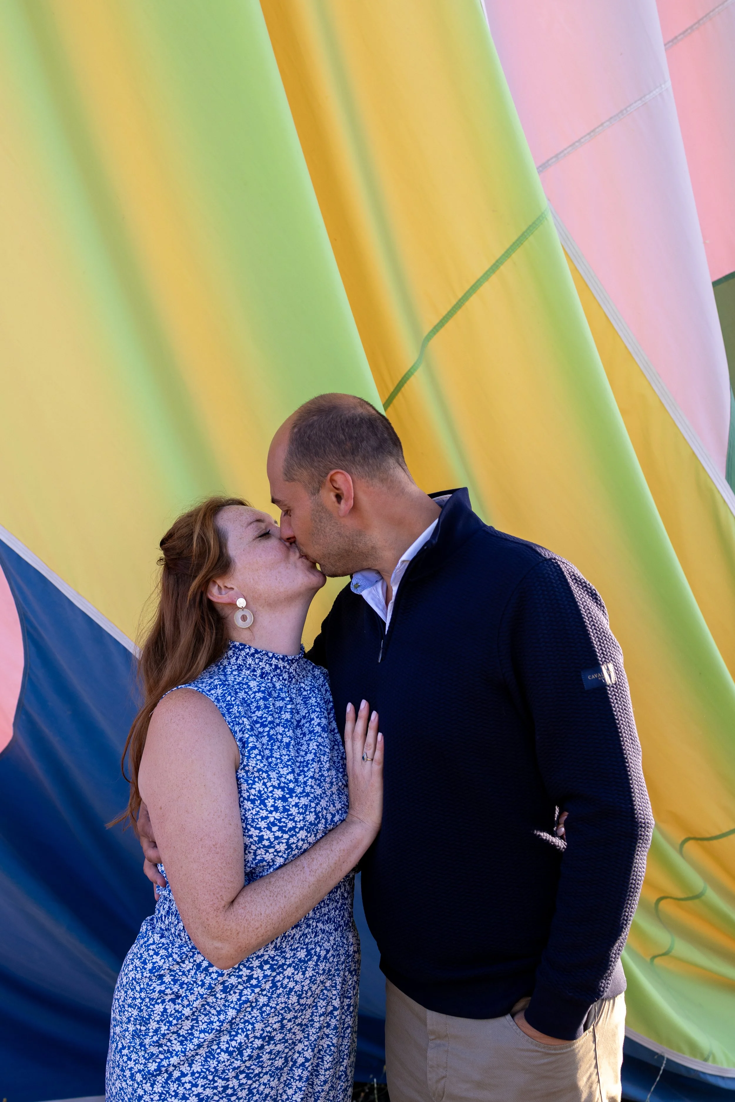A couple kissing in front of a colorful hot air balloon.