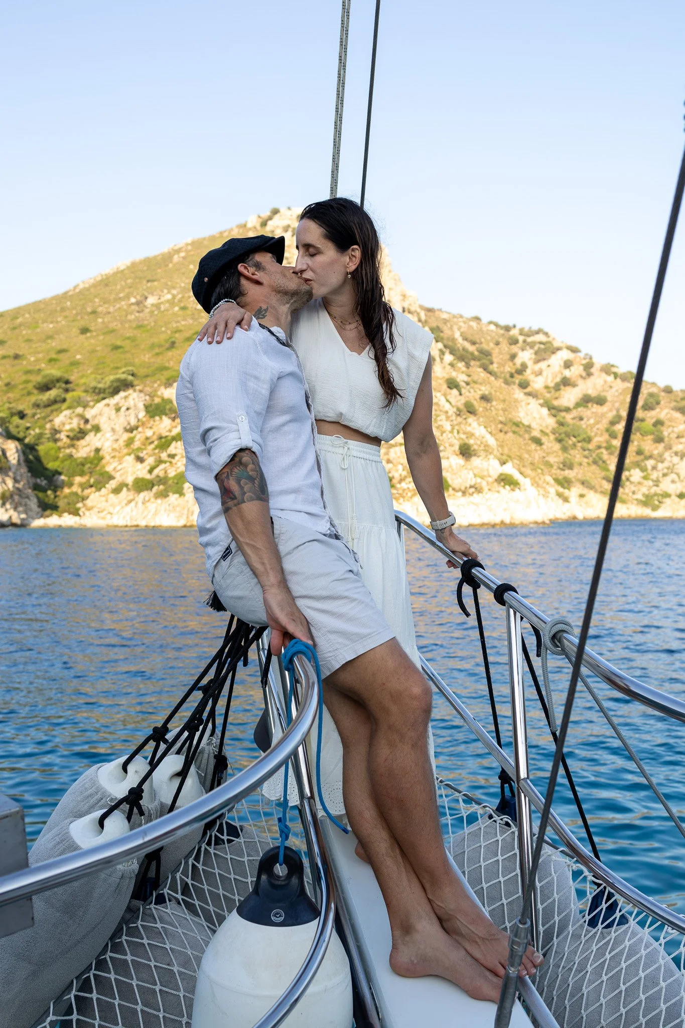 A couple sharing a kiss on a sailboat with a water and hillside background.