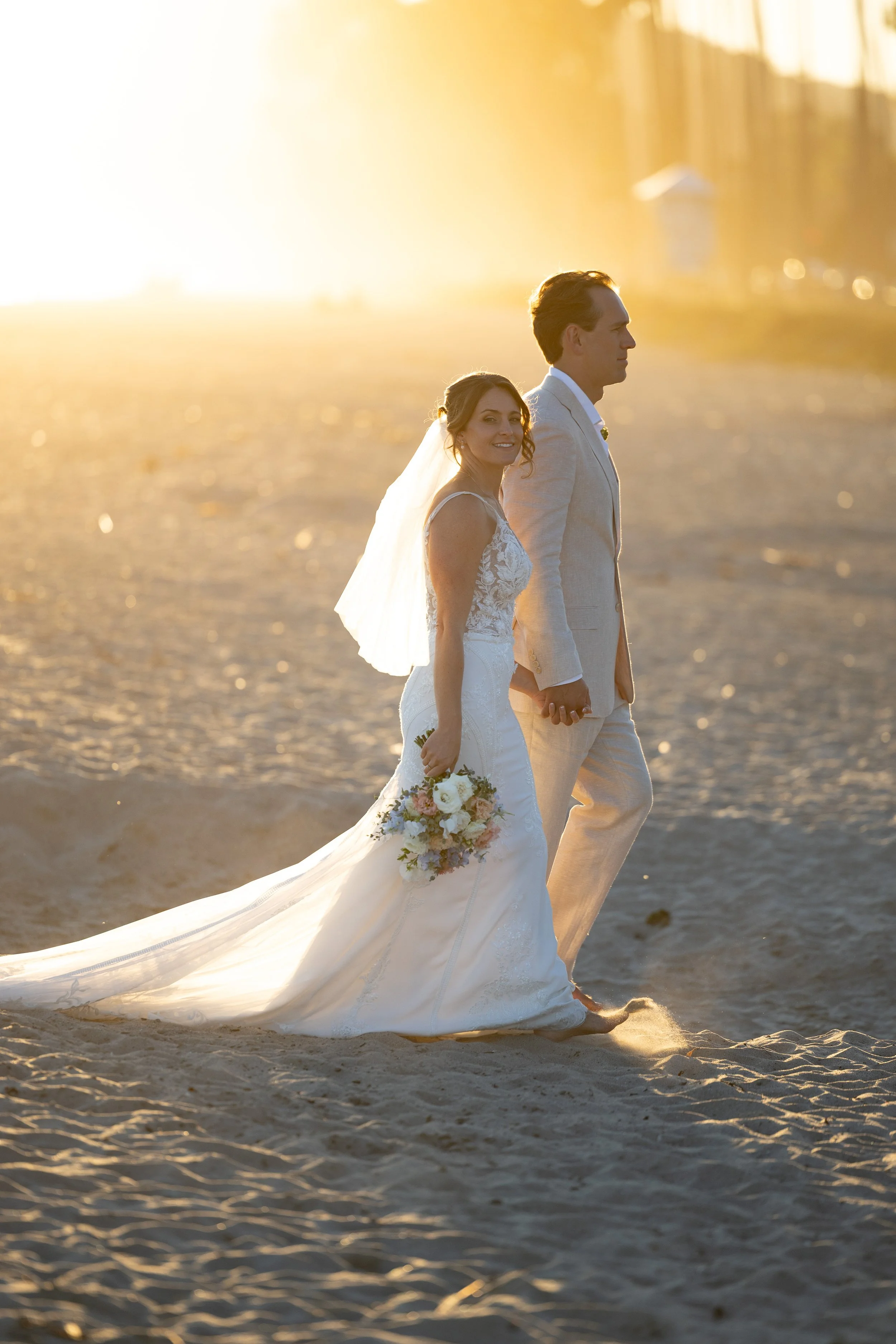 A bride and groom walking hand in hand on the beach during sunset, with the bride holding a bouquet of flowers and wearing a white wedding dress and veil, and the groom dressed in a light-colored suit.