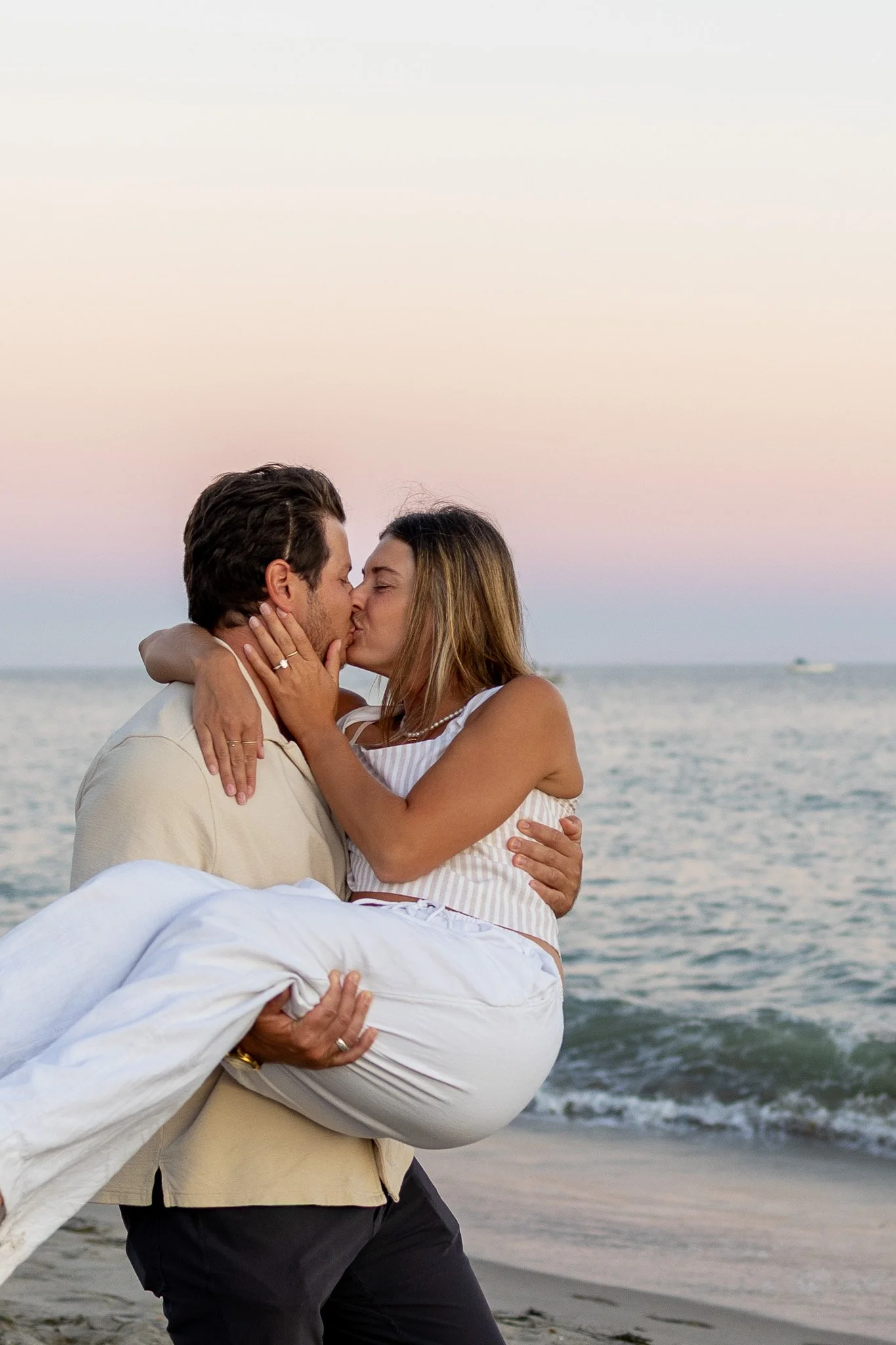 A couple kissing on the beach during sunset, with the man holding the woman in his arms and audience of the ocean in the background.