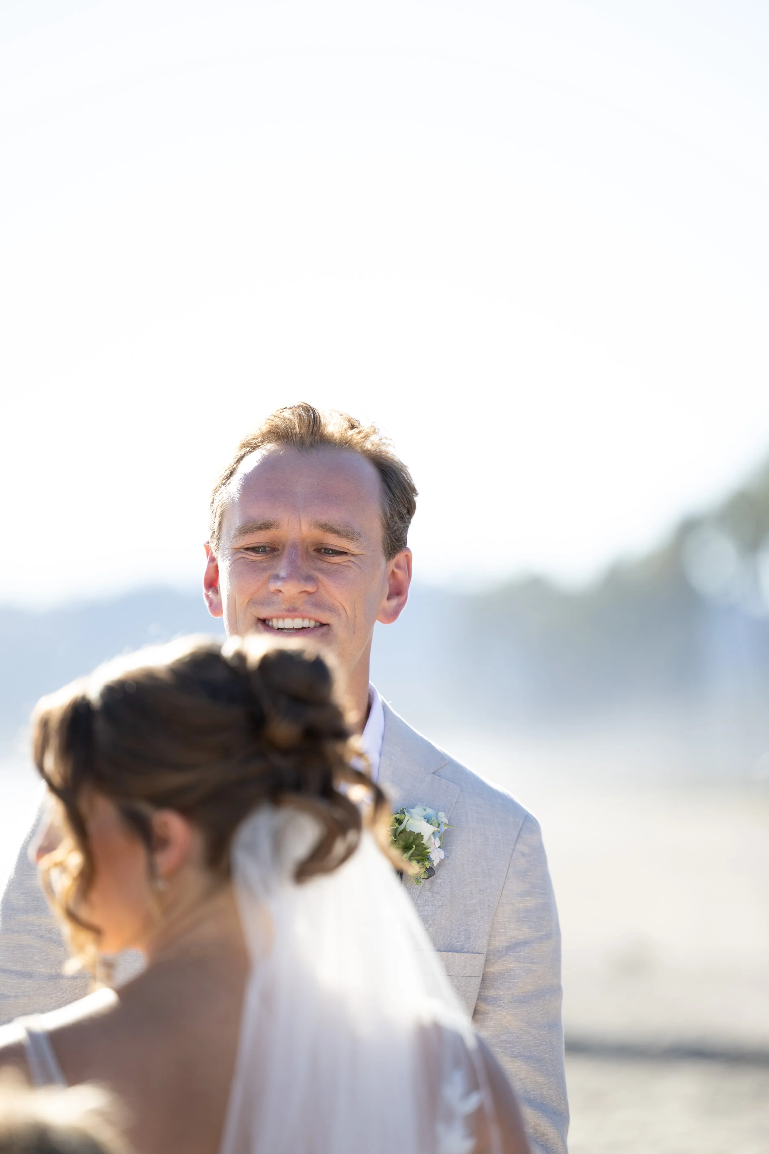 A man in a light gray suit smiling at a woman in a wedding dress, outdoors on a bright sunny day with a blurred background.
