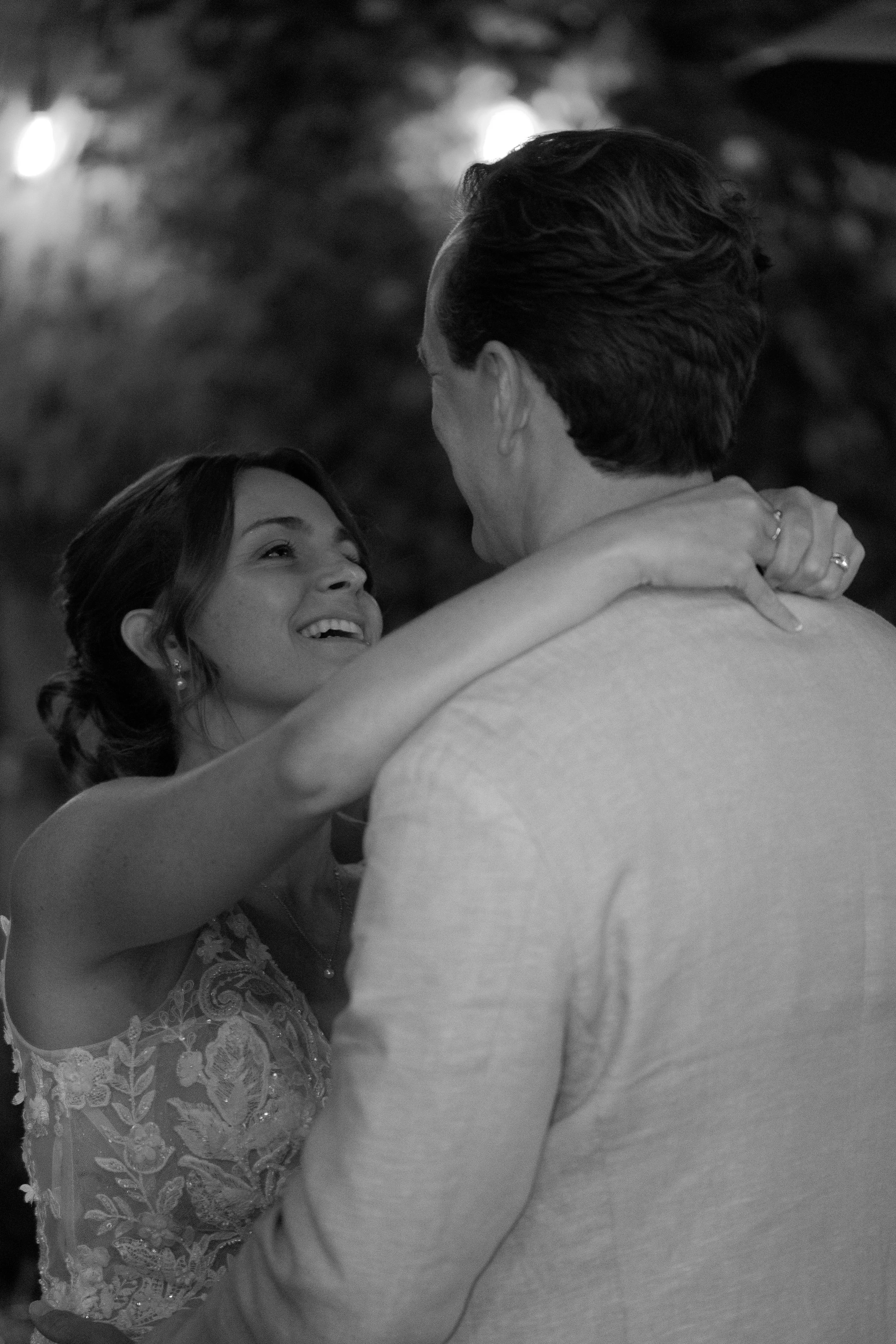 A woman and man are dancing closely, looking into each other's eyes with smiles. The woman has her arms around the man's neck, and the man faces her with his back to the camera. It is a joyful, intimate moment.