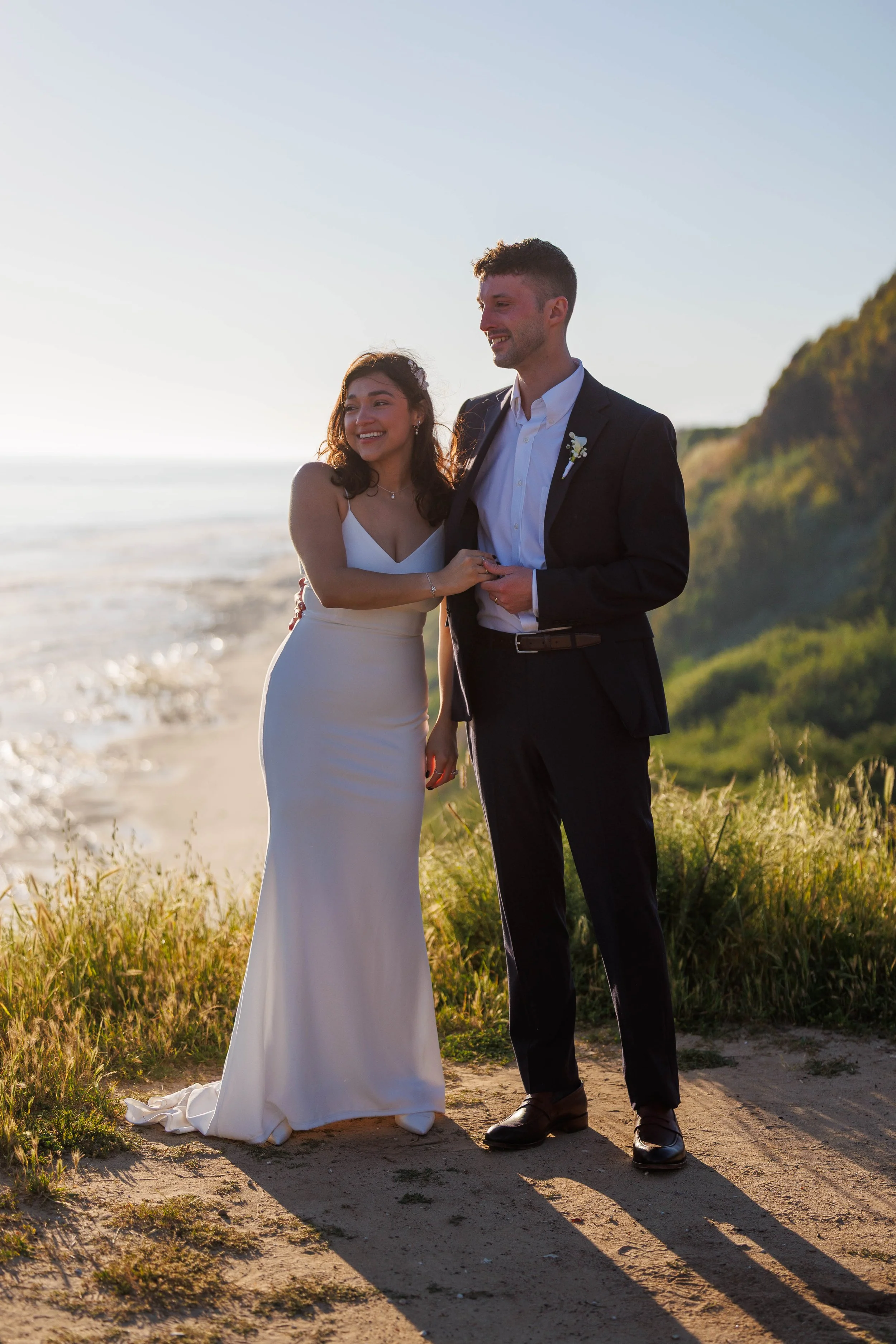 A bride and groom smiling on their wedding day at the beach during sunset, holding hands and standing on sandy ground with grass and a backdrop of ocean and hills.