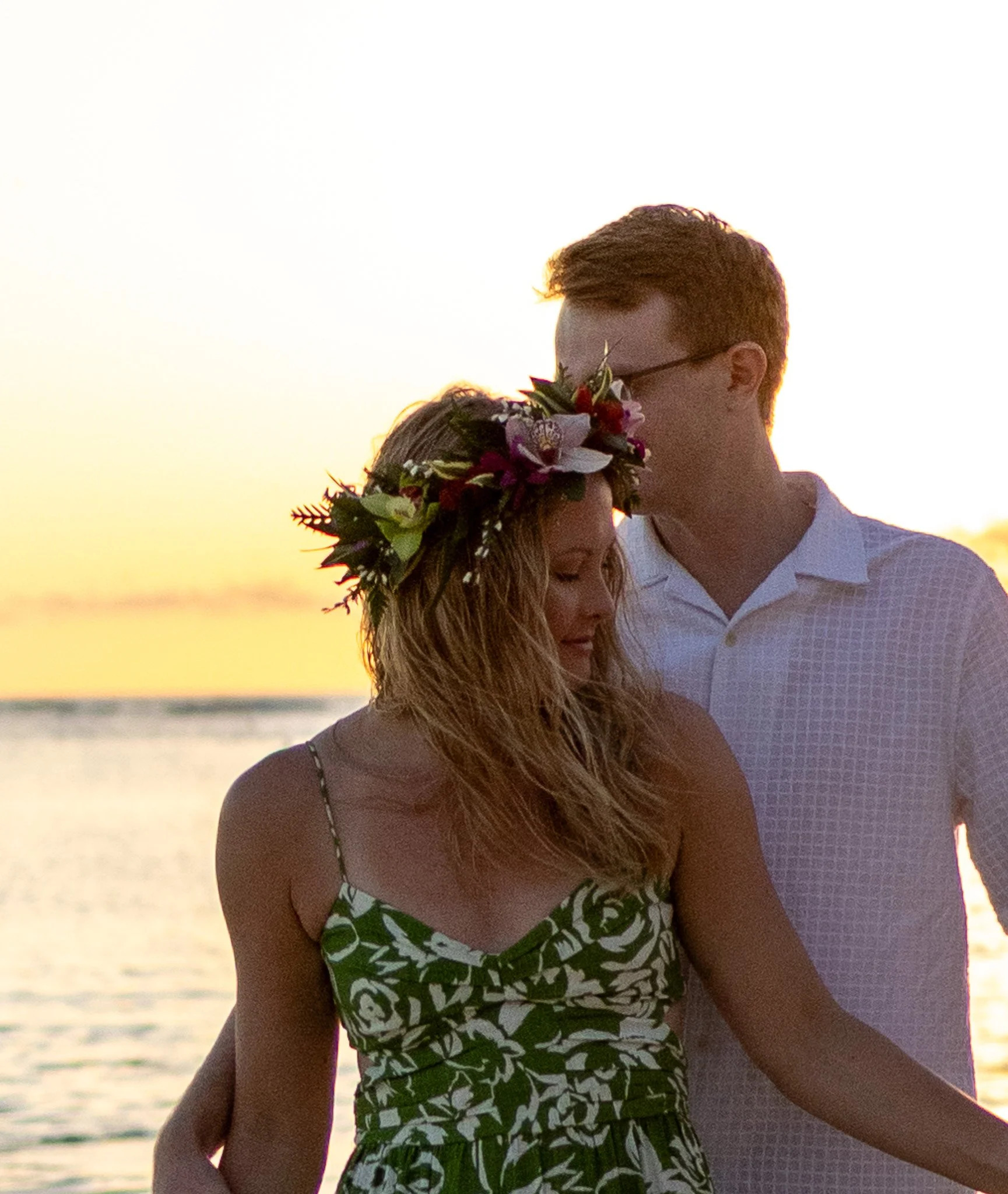 A couple standing close together at sunset by the water, with the man leaning in to kiss the woman's forehead, wearing a floral crown and summer dress.