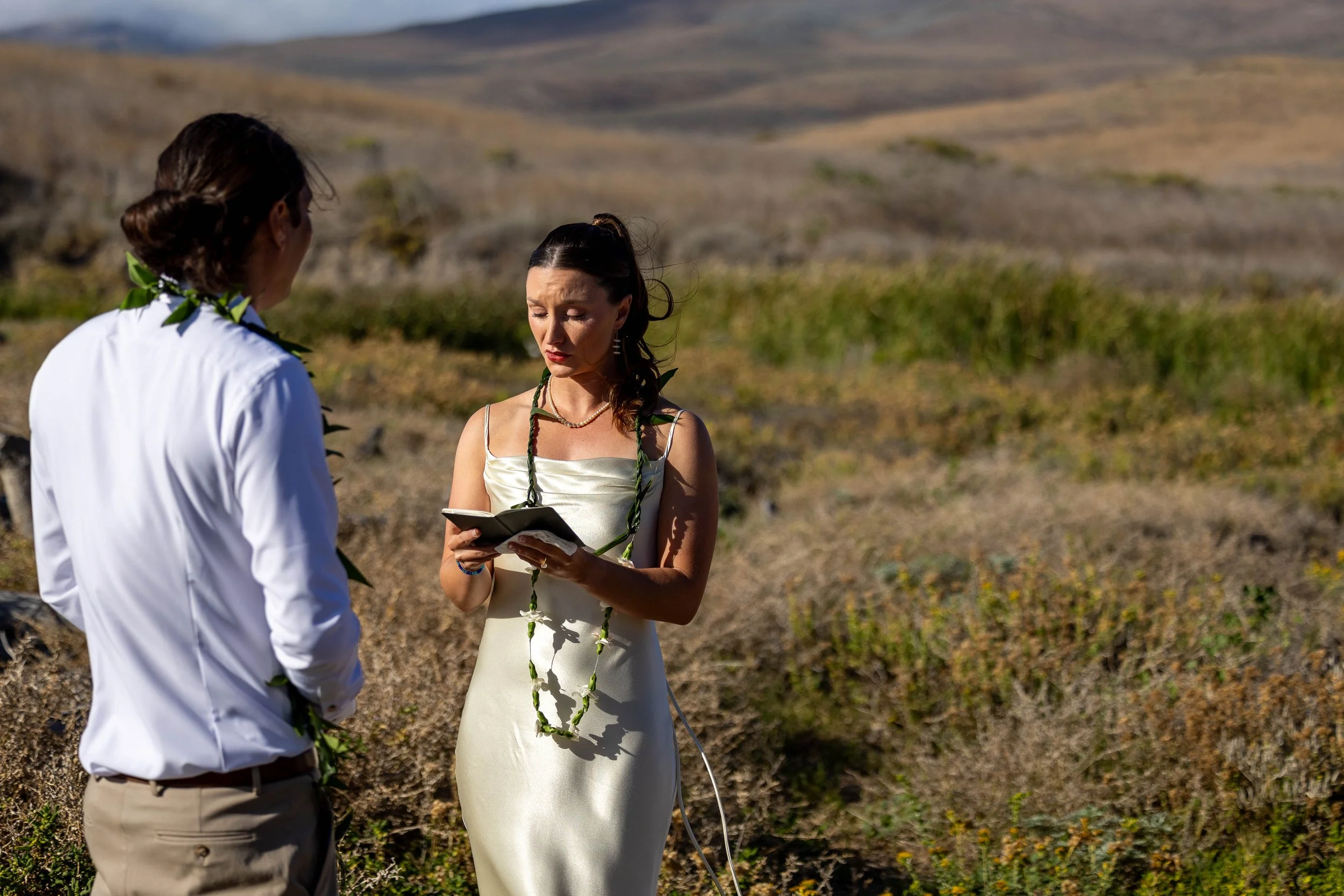 A couple dressed in wedding attire standing outdoors in a dry, hilly landscape, with the woman reading from a book or paper.