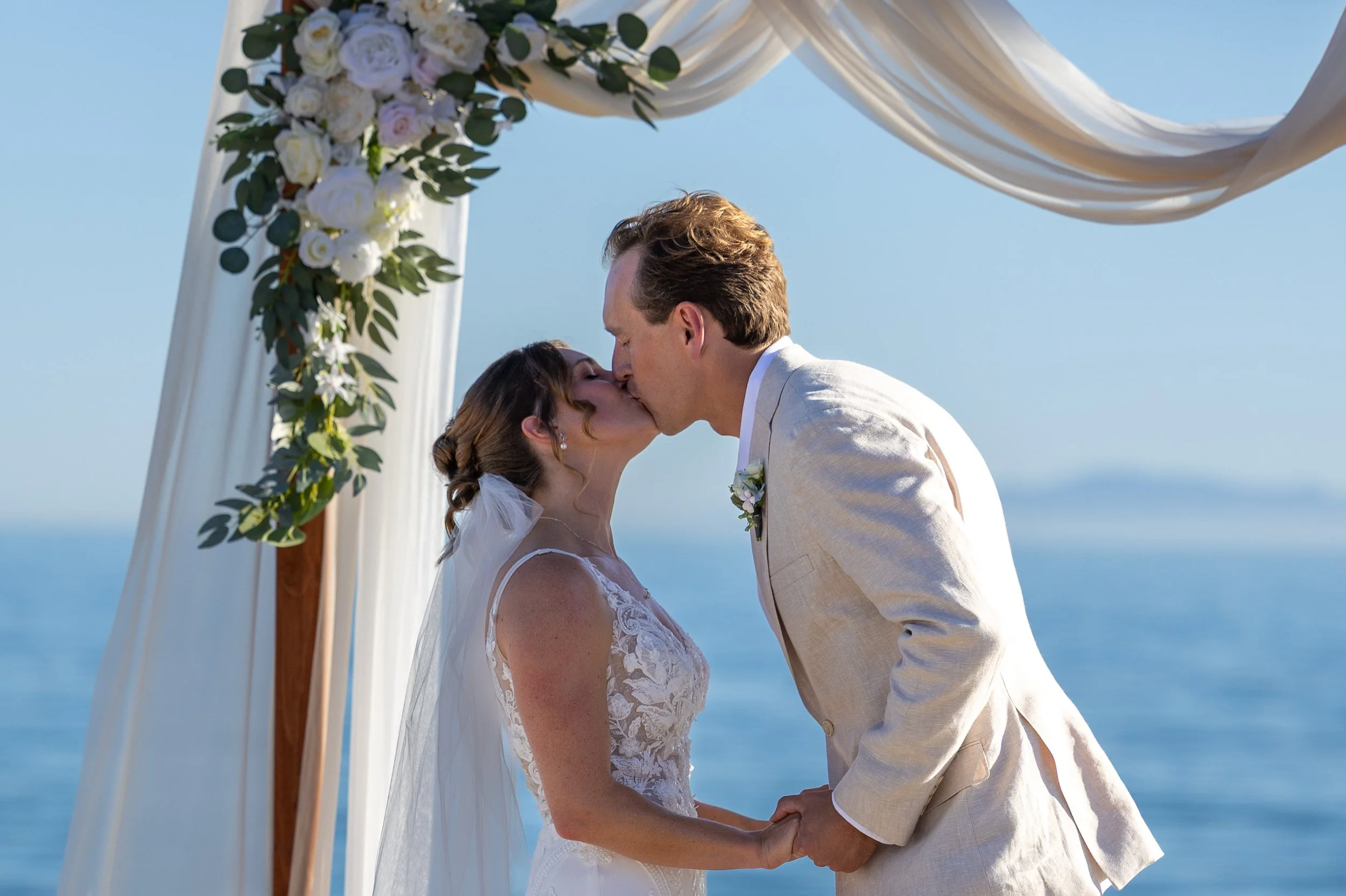 A bride and groom share a kiss under a decorated wedding arch by the ocean.