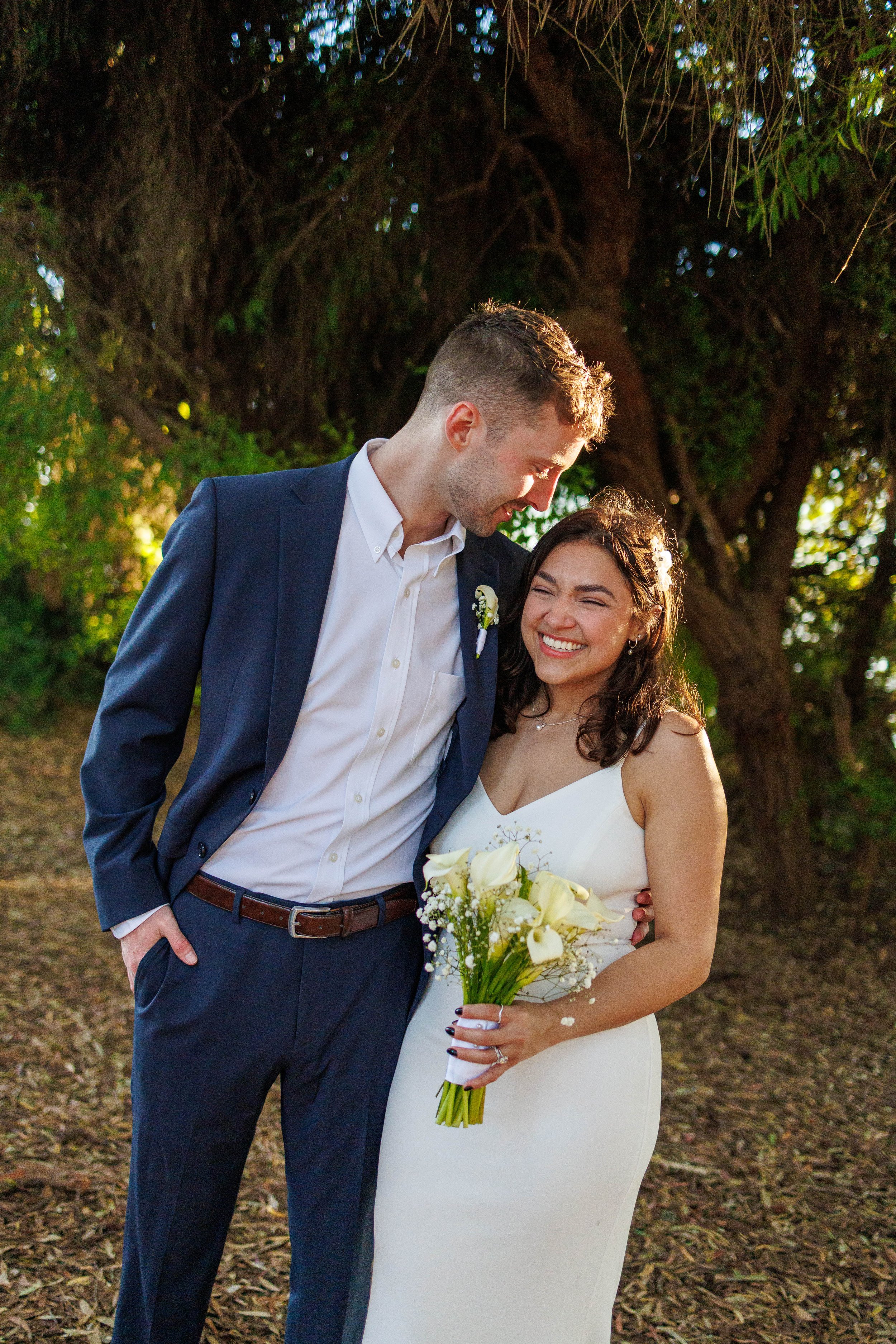 A bride and groom share a joyful moment outdoors, with the man leaning his forehead against the woman's head. The bride is holding a bouquet of white flowers and is smiling widely, while the groom is looking affectionately at her. They are surrounded