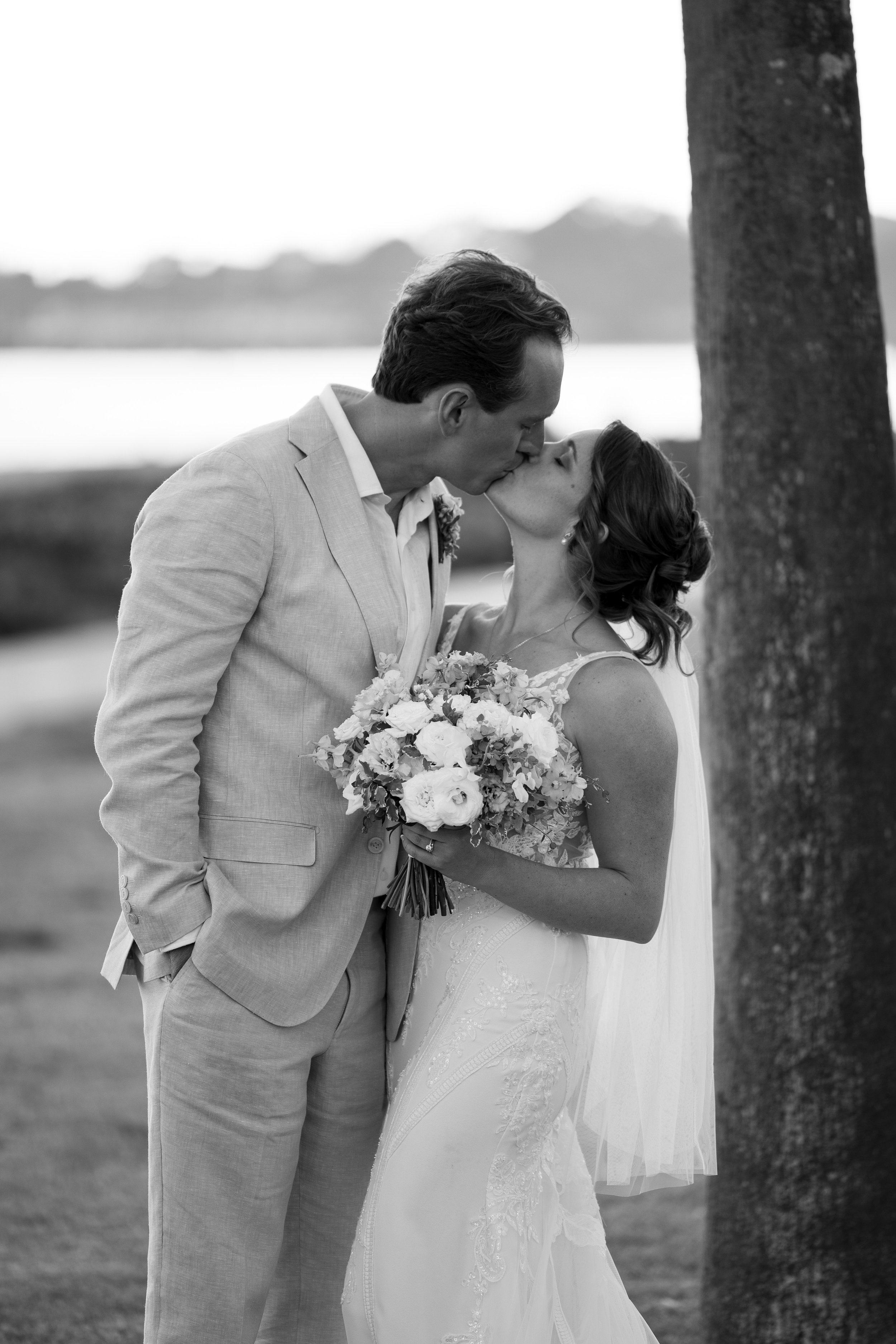 A black and white photo of a bride and groom kissing outdoors on their wedding day, standing near a tree with a body of water and trees in the background.