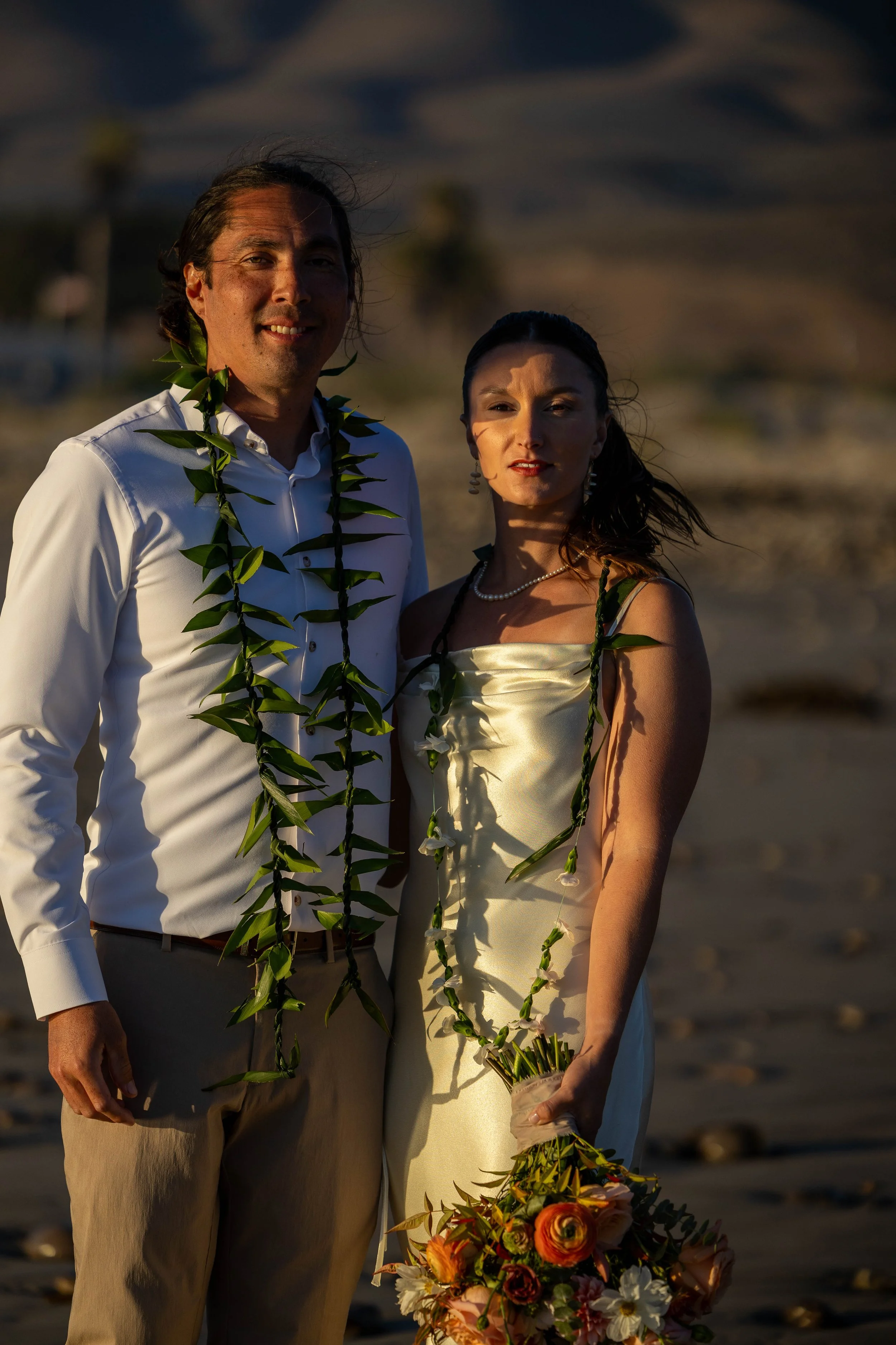 A couple dressed in wedding attire standing on a beach during sunset, holding a bouquet of flowers, with greenery leis around their necks.
