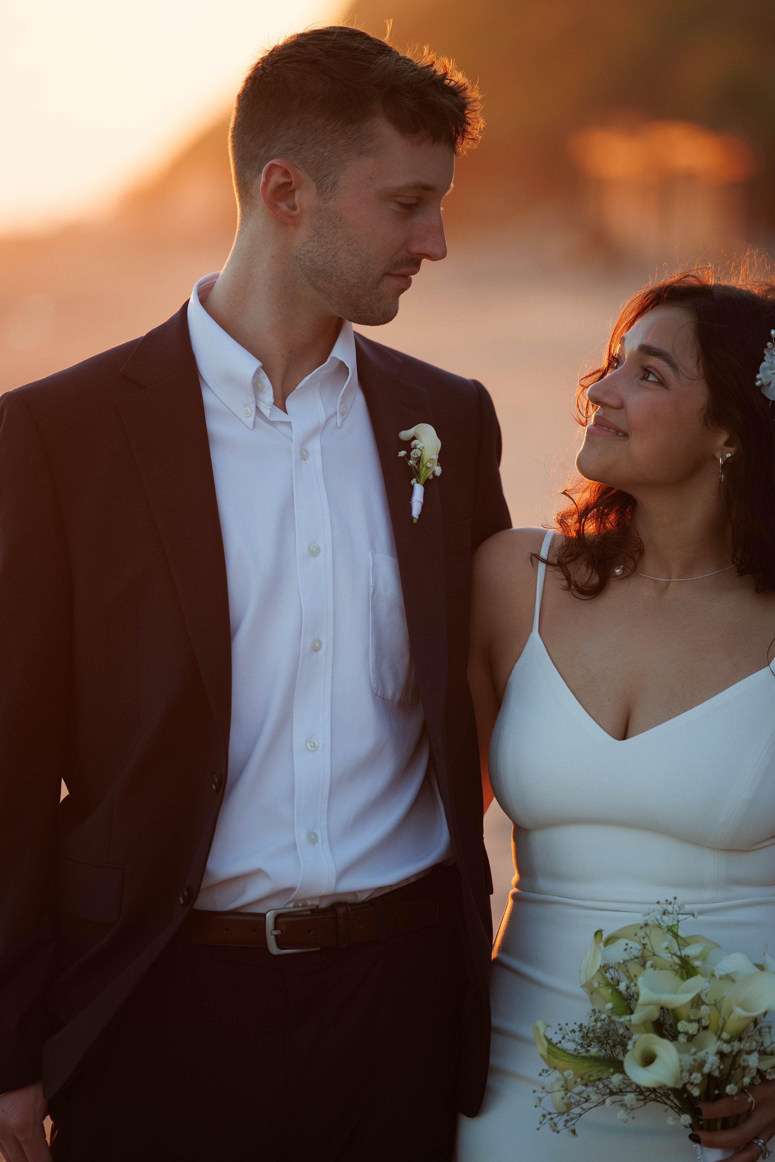 A bride and groom standing close together outdoors at sunset, with the groom wearing a black suit and white shirt, and the bride in a white dress holding a bouquet of white flowers, both smiling at each other.
