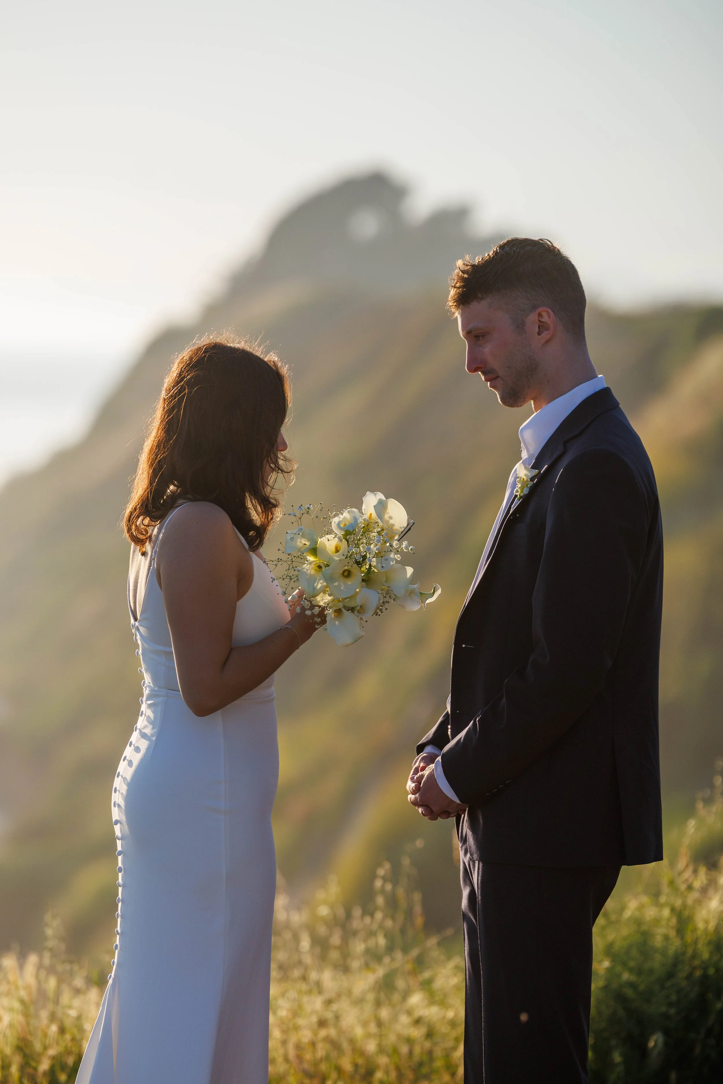 A bride and groom standing outdoors on a hill at sunset, facing each other with the bride holding a bouquet of white flowers, with a scenic background of cliffs and sky.