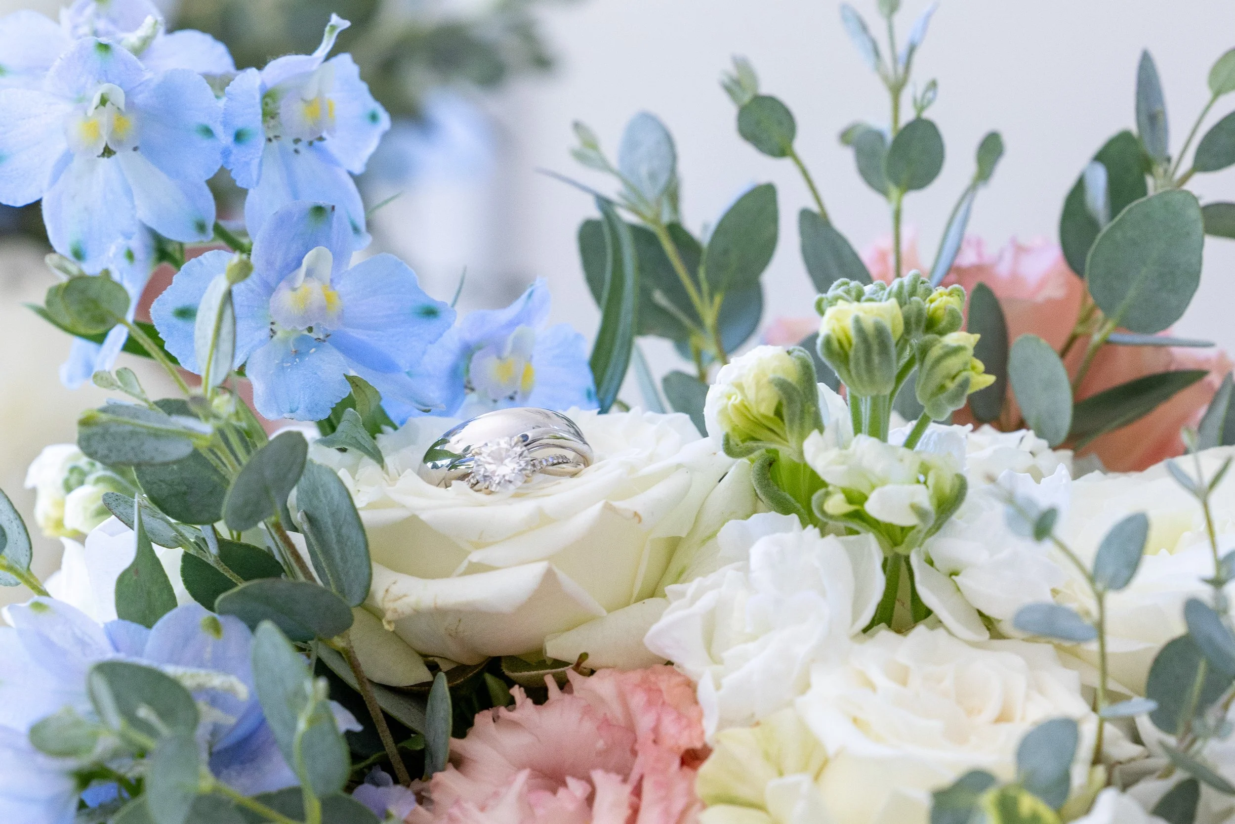 Close-up of a floral arrangement with white roses, blue delphiniums, pink carnations, and green foliage. A diamond engagement ring rests on a white rose in the center.