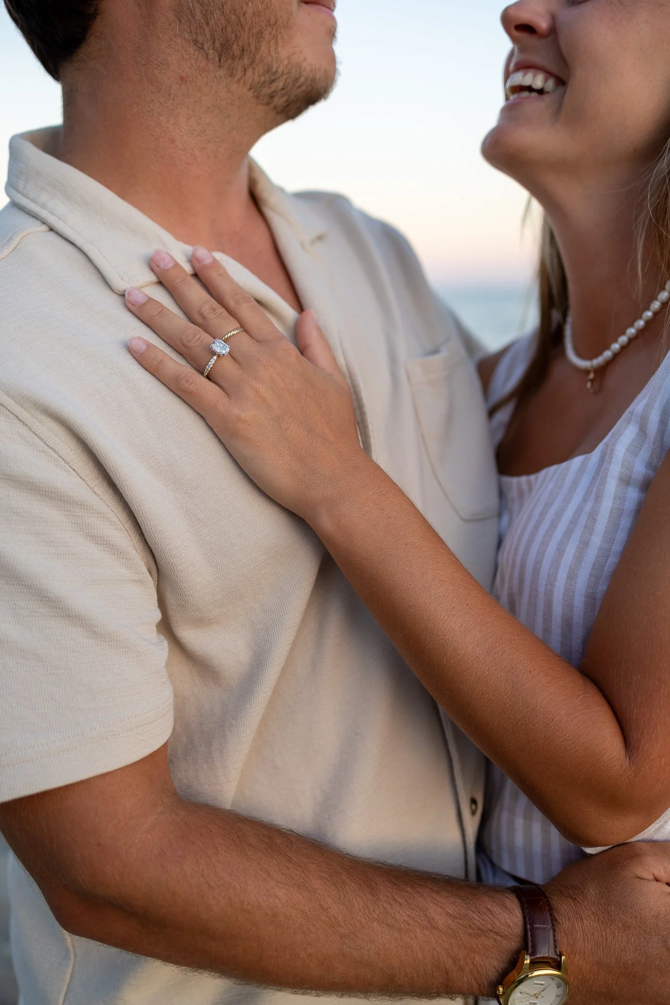 Close-up of a couple embracing, with a focus on the woman's hand displaying a diamond engagement ring, smiling at each other in an outdoor setting.