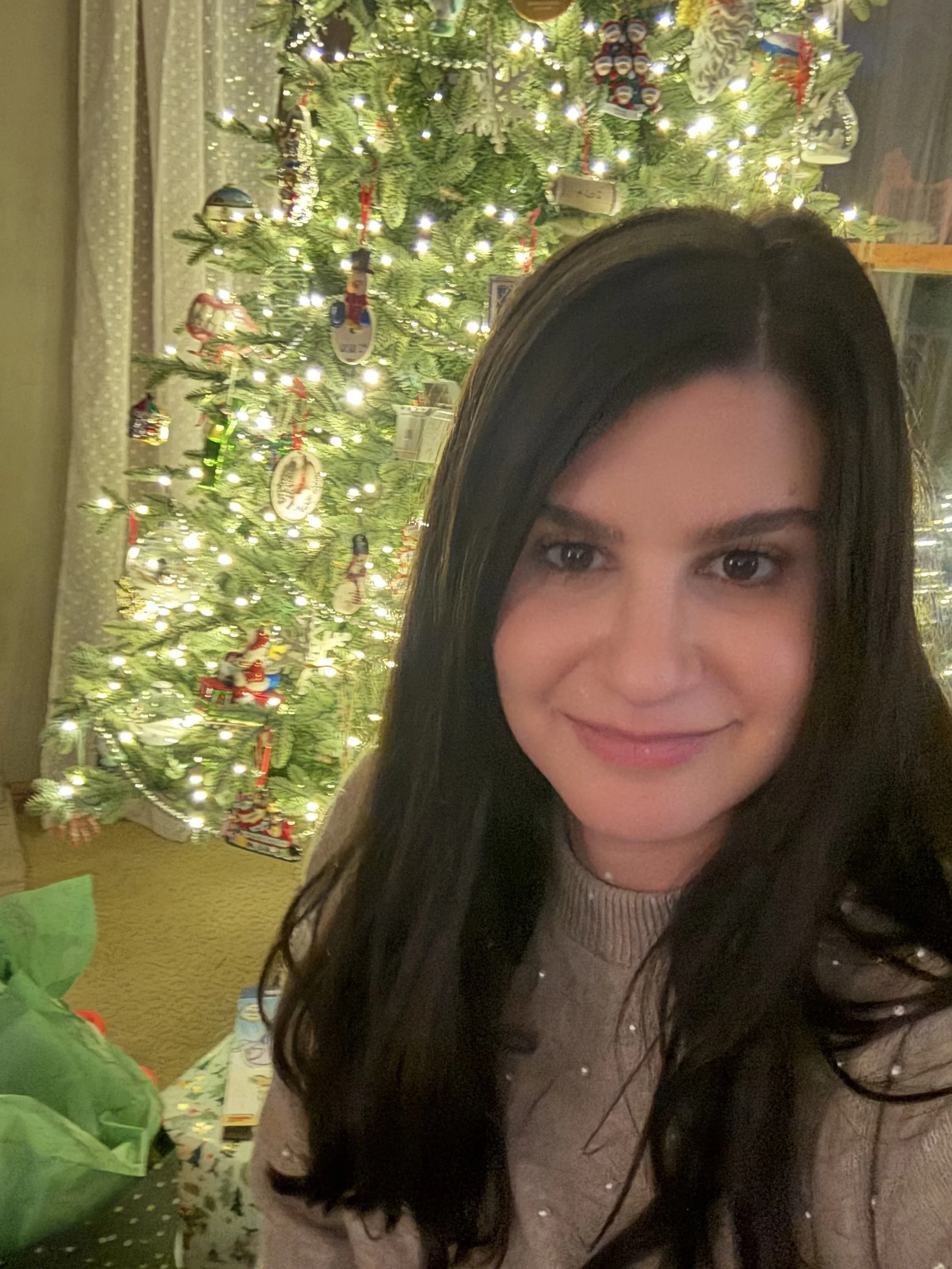 A woman with long dark hair smiling in front of a decorated Christmas tree with lights and ornaments.