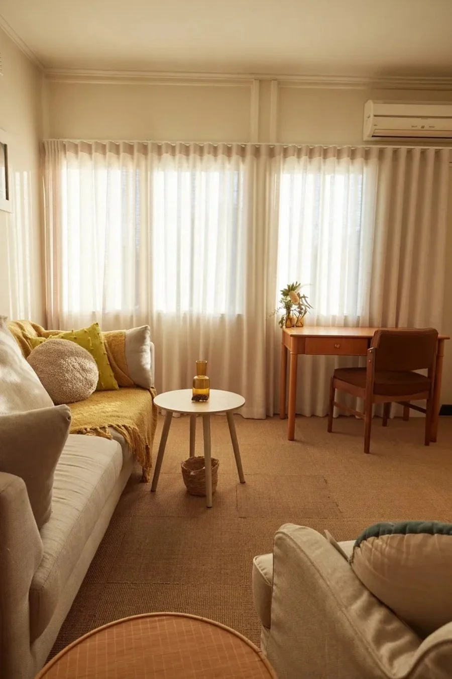The consulting room: soft daylight through sheer curtains, a sofa with cushions, a small side table and a timber desk