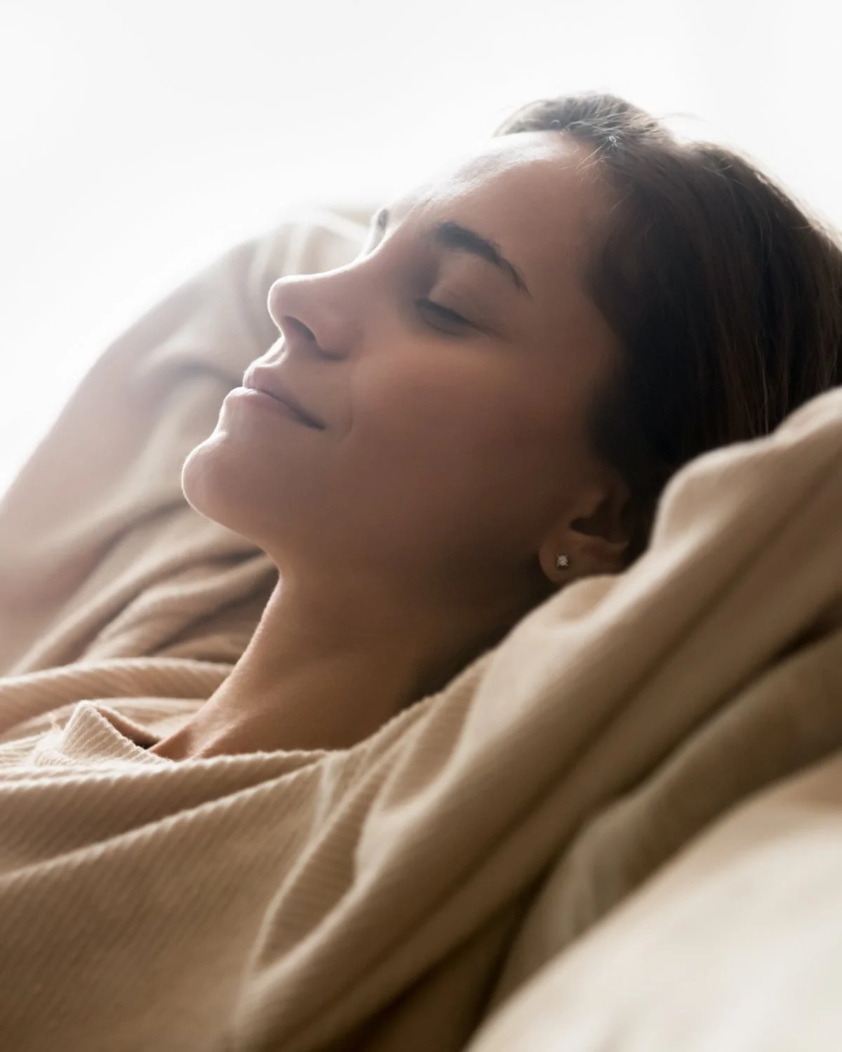 A woman resting with her eyes closed, wrapped in a soft blanket in gentle morning light